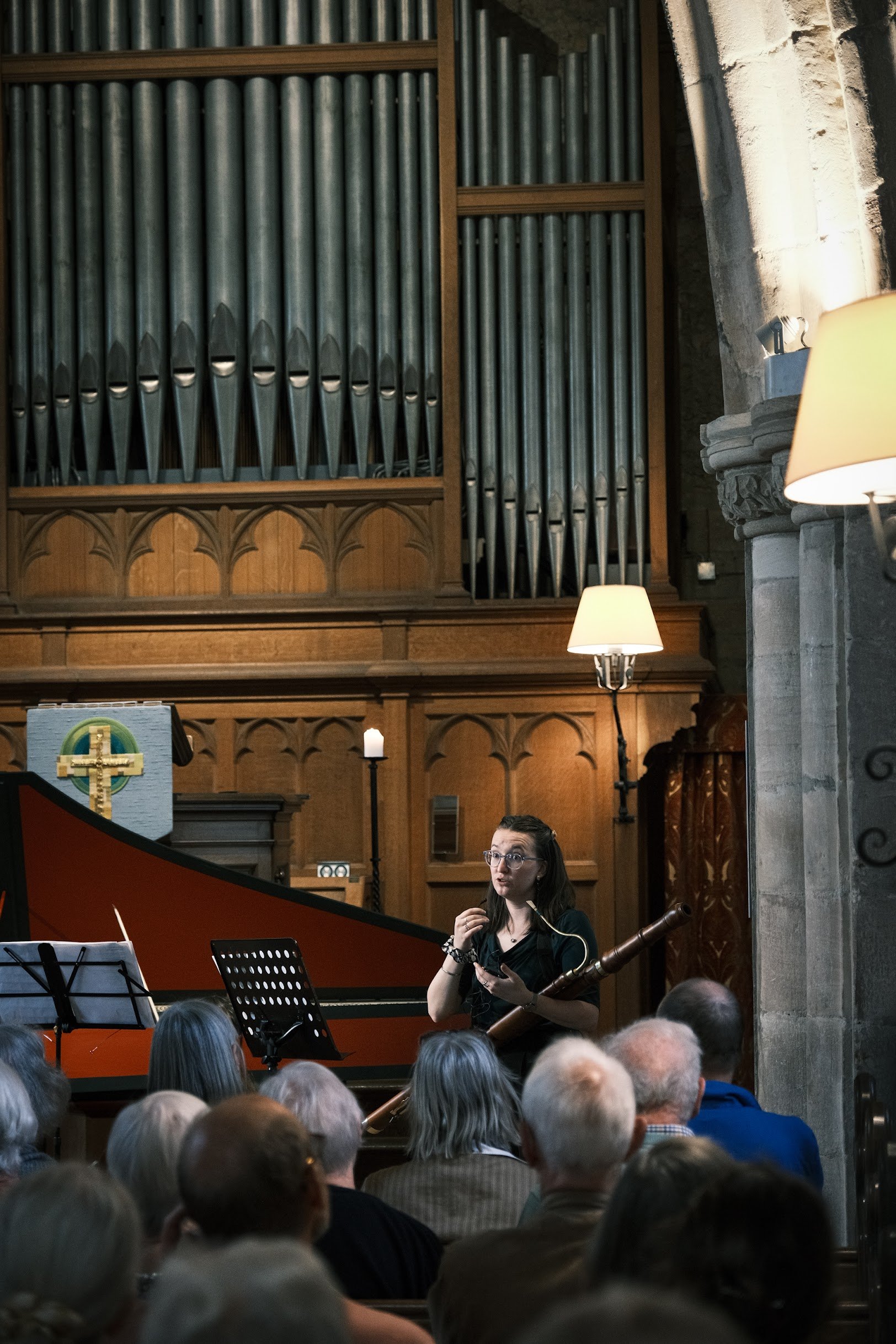 A woman holding a baroque bassoon, speaking to an audience inside a church with wooden paneling, large organ pipes, and lit lamps on the walls.
