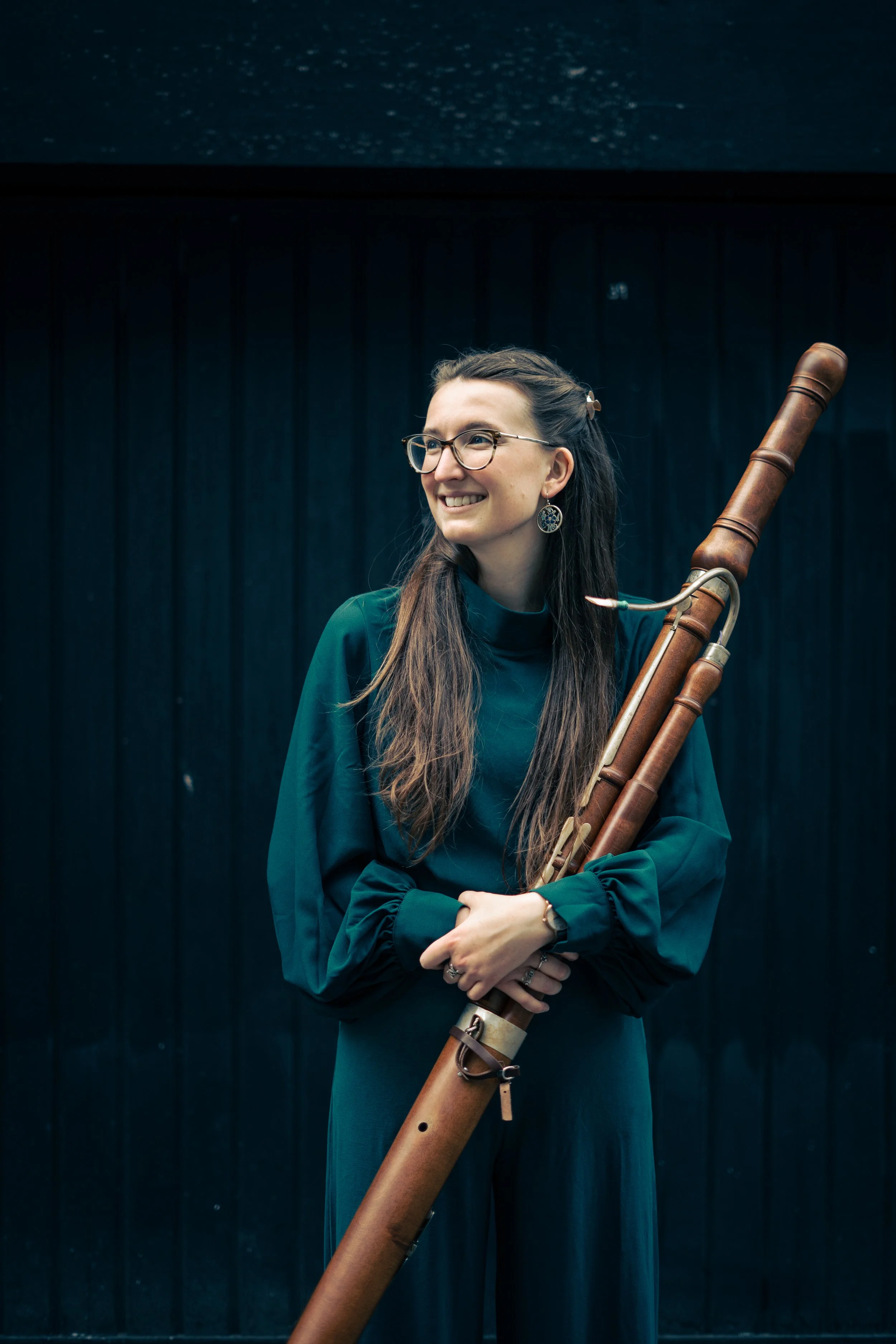 A woman with long brown hair, glasses and earrings, dressed in dark green, holds a baroque bassoon, standing against a dark background.