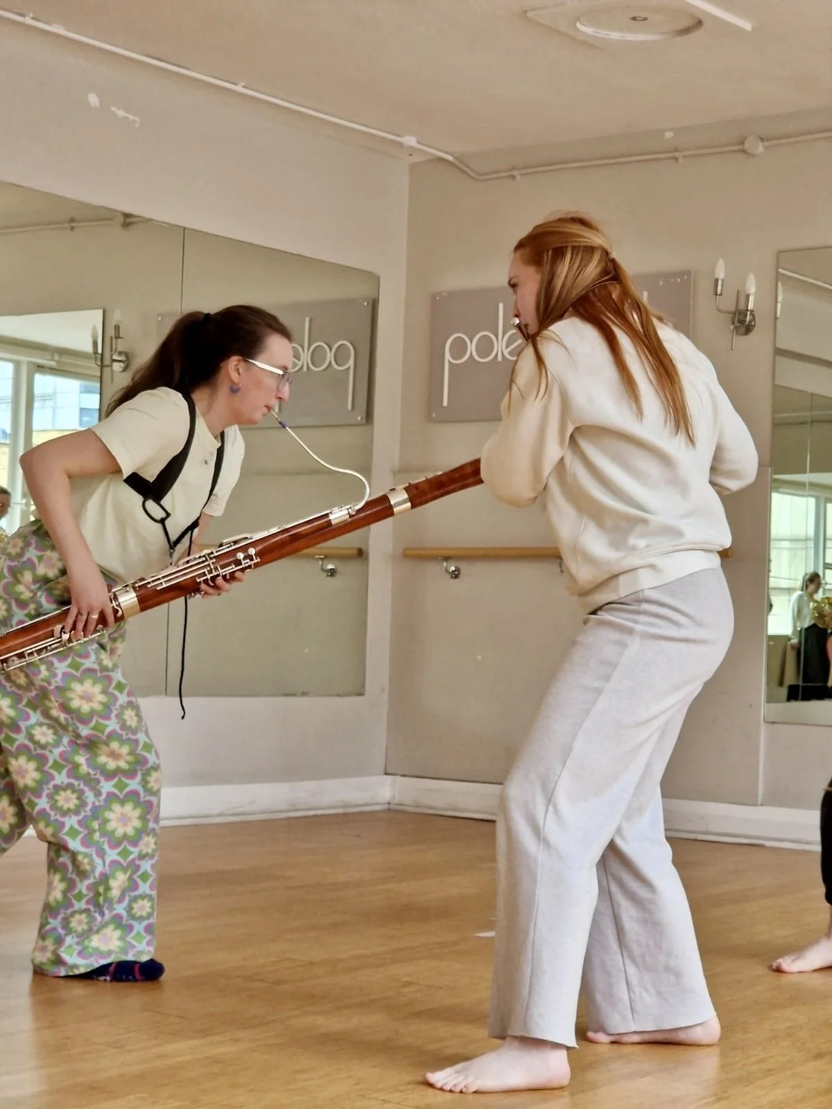 Two women leaning towards each other, one playing a bassoon and the other playing a flute, in a dance studio.
