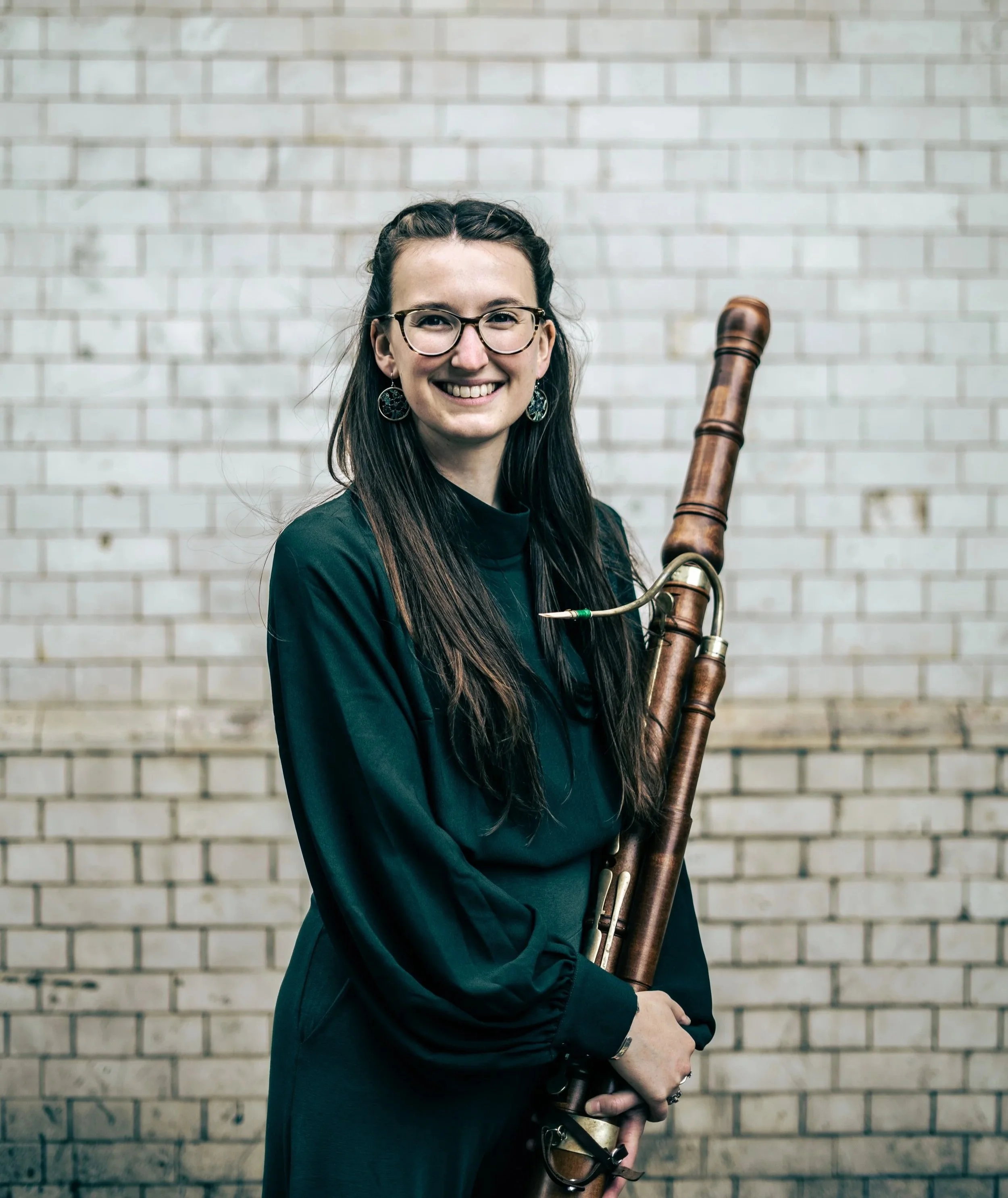 A smiling woman with long dark hair, glasses, and earrings, dressed in green, holding a baroque bassoon against a white brick wall.