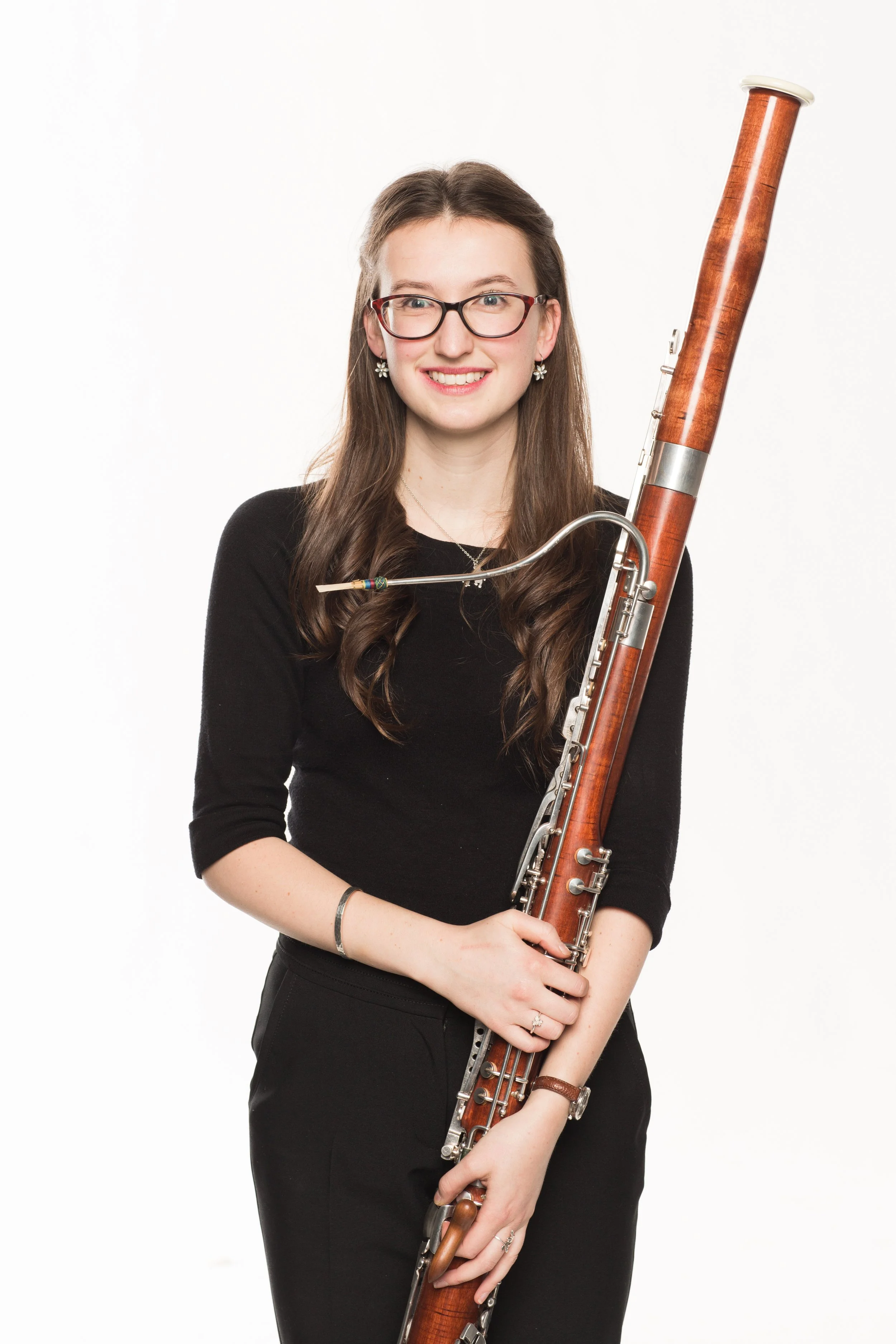 Young woman with long brown hair, wearing glasses and dressed in black, holding a bassoon and smiling in front of a plain white background.