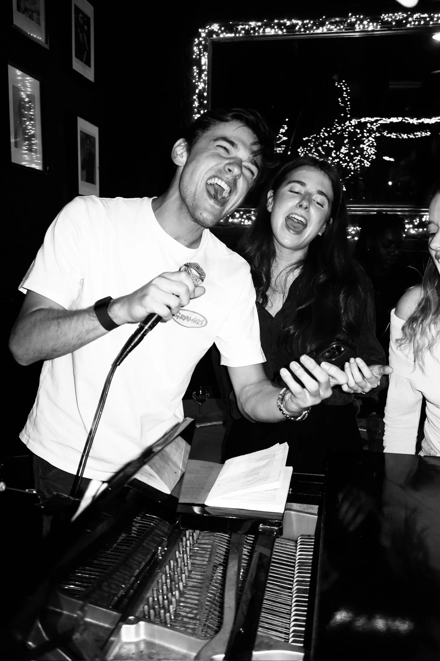 Two people singing or performing, one holding a microphone, in a lively indoor setting with festive string lights.