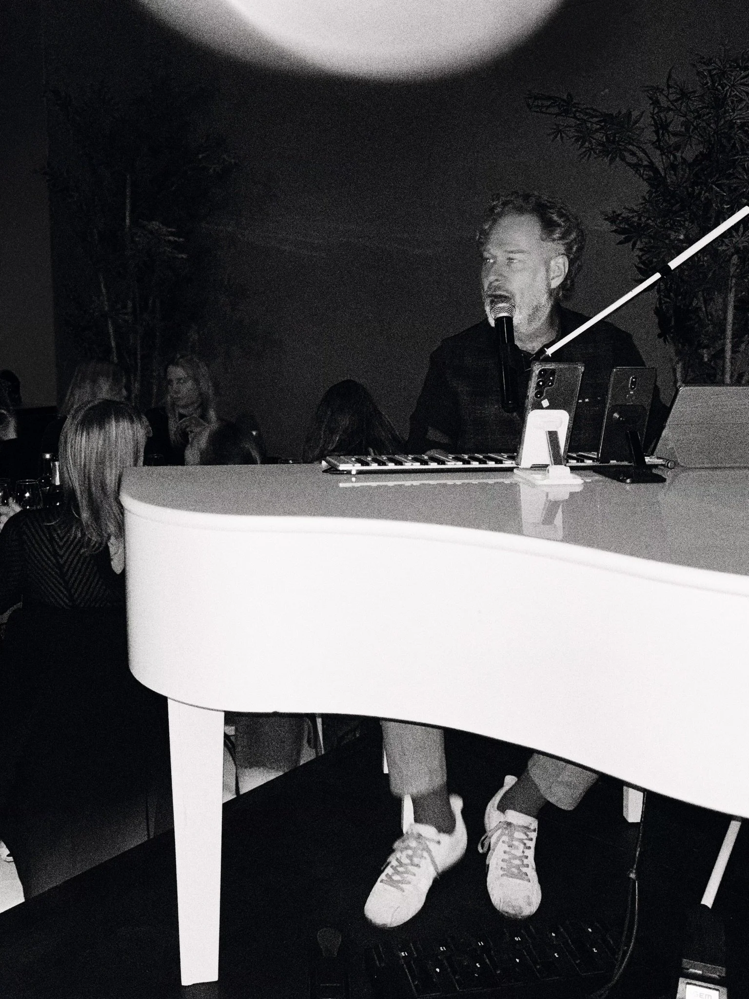 A man singing into a microphone at a piano, with several smartphones placed on the piano's surface, in a dimly lit room filled with people, some seated and others standing, and illuminated by a circular light fixture overhead.