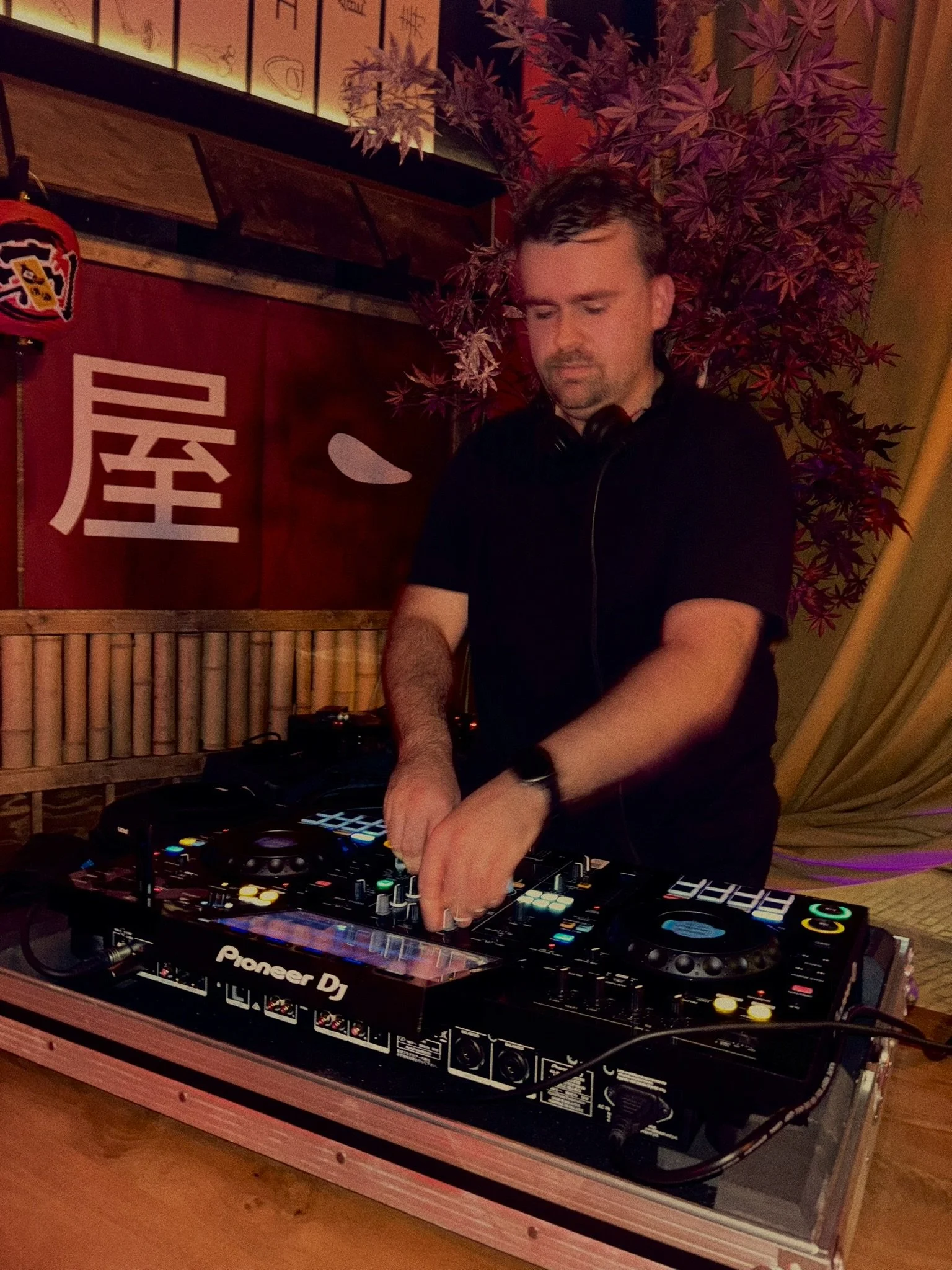 A man DJing at a club or bar, using a Pioneer DJ controller. He wears a black shirt and has headphones around his neck, with a decorative Japanese-style wall and purple foliage in the background.