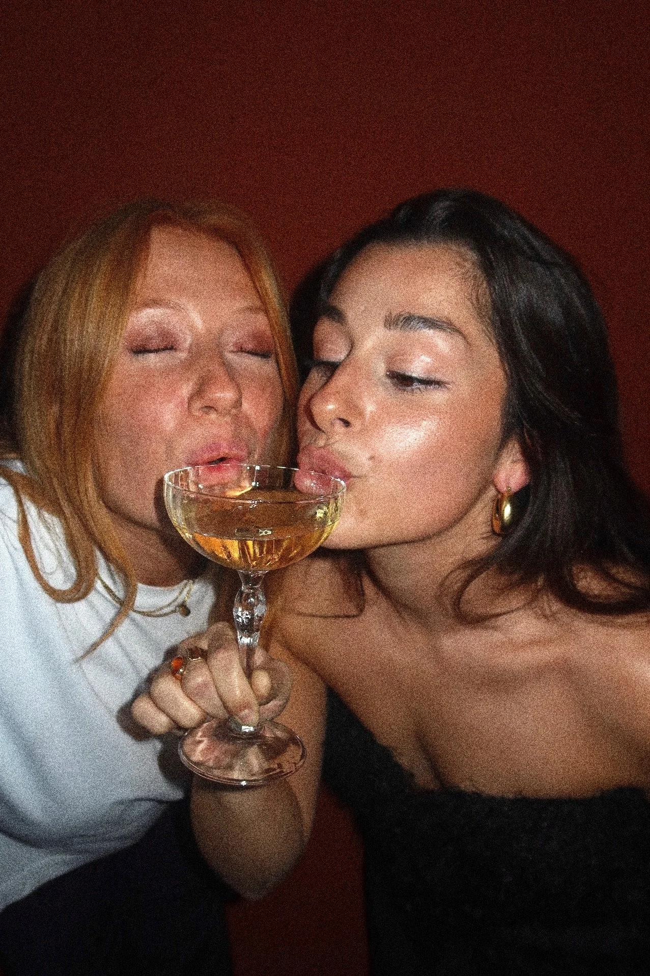 Two women are close together, one with red hair and the other with dark hair, sharing a drink in a cocktail glass. They have their eyes closed and are smiling.