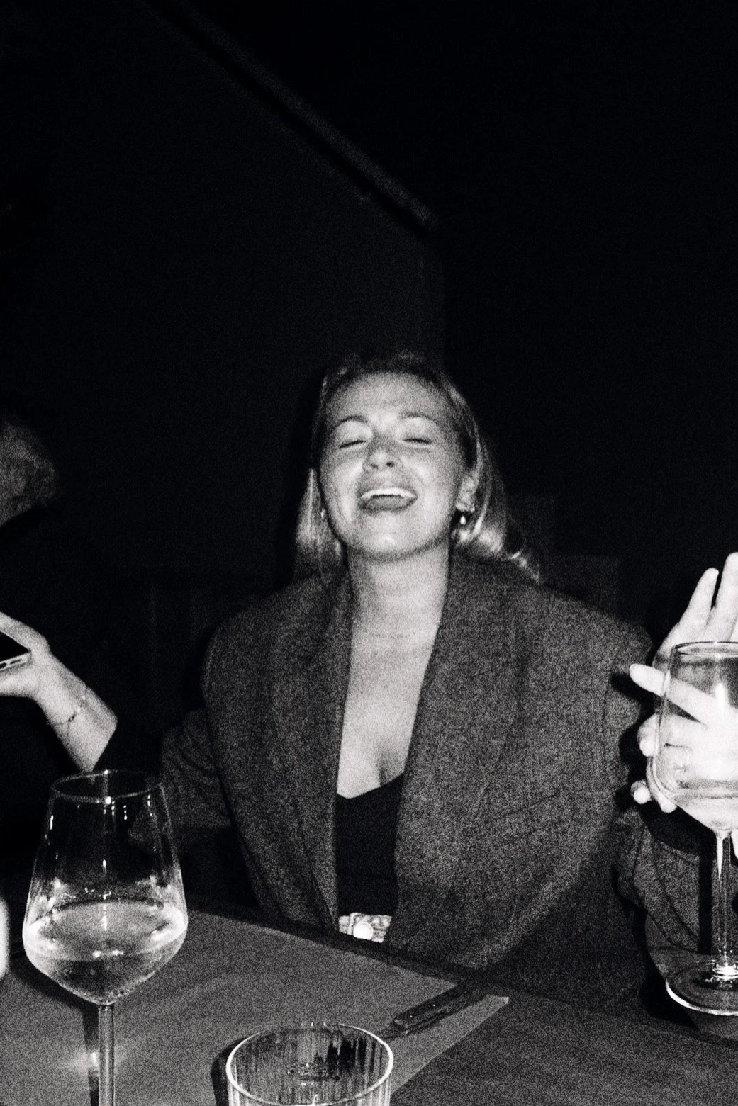 A woman with short, blonde hair smiling and laughing at a dinner table, holding a glass of wine with her left hand, in a dark indoor setting.