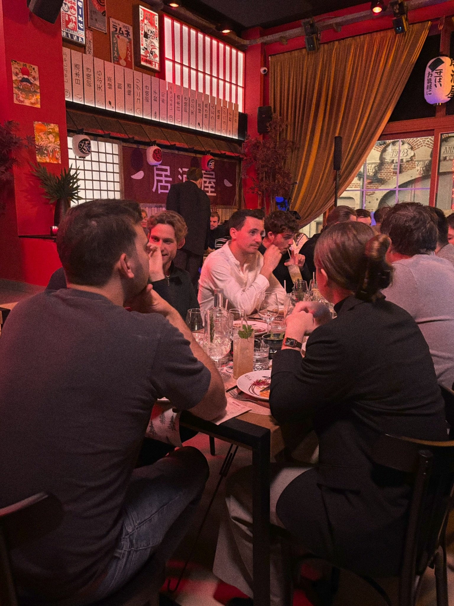 People seated around a table in a Japanese-themed restaurant with red walls, Japanese banners, and paper lanterns. There are drinks and plates of food on the table, and a curtain and window with a view outside.
