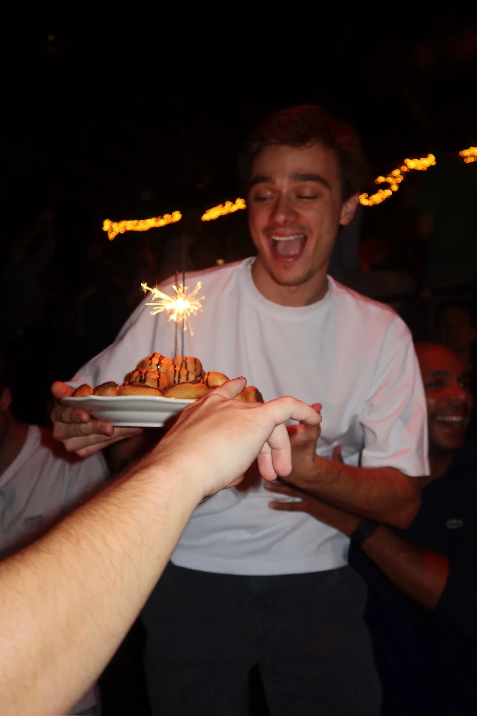 A young man holding a plate of desserts with a sparkler on top, smiling excitedly during a celebration at a party or restaurant.