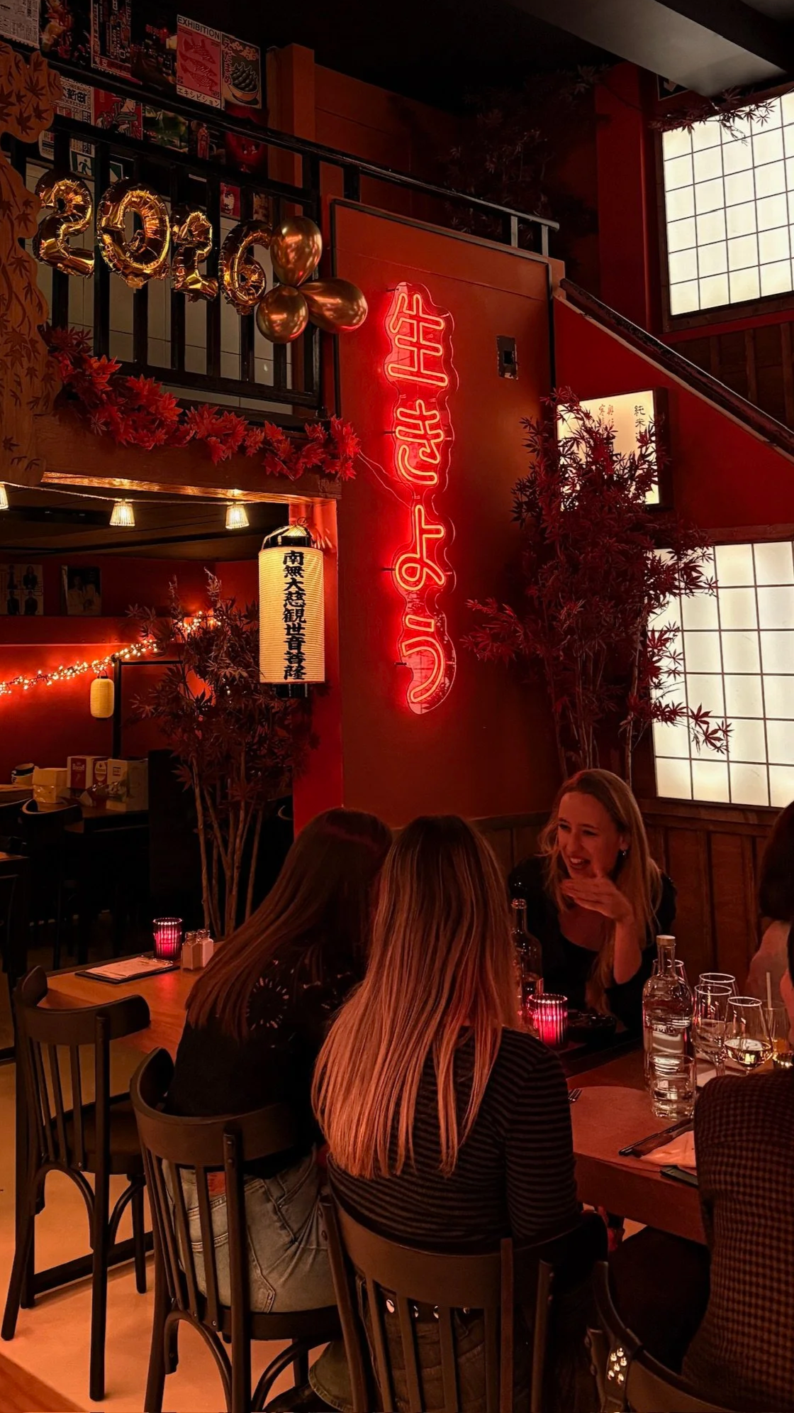 Interior of a restaurant decorated with Halloween and New Year 2016 decorations, including balloons and a neon sign with Chinese characters, with women sitting at a table talking and smiling.