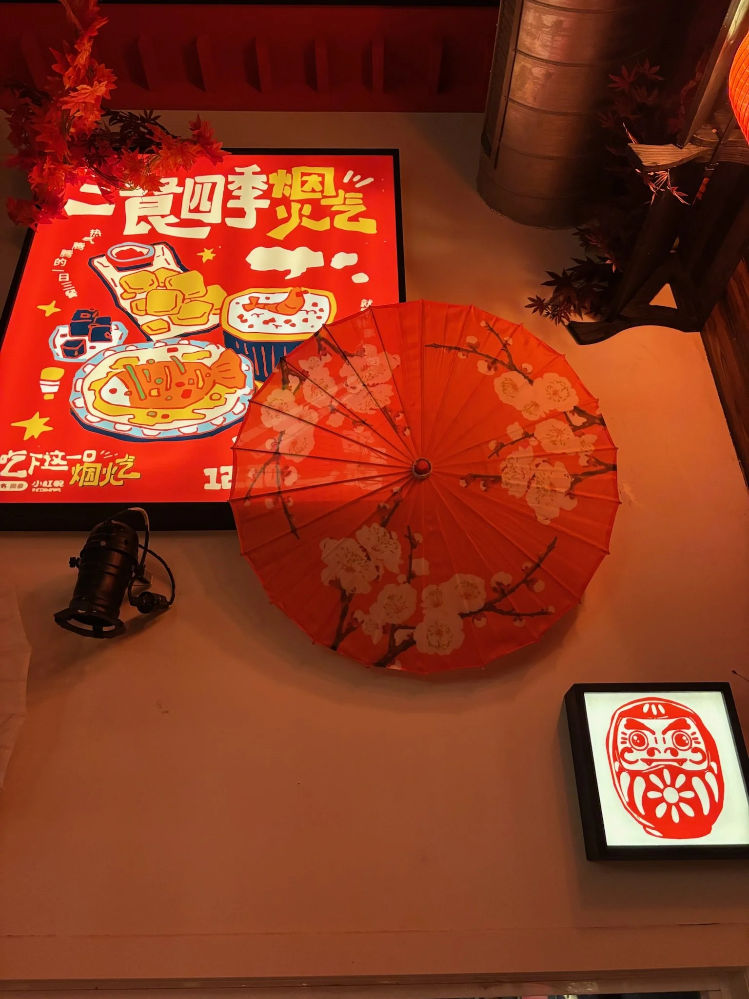 Decorative Chinese-themed display with a red paper parasol with floral patterns, a bright red sign featuring food illustrations and Chinese characters, a small Daruma doll image, and a spotlight on the floor.