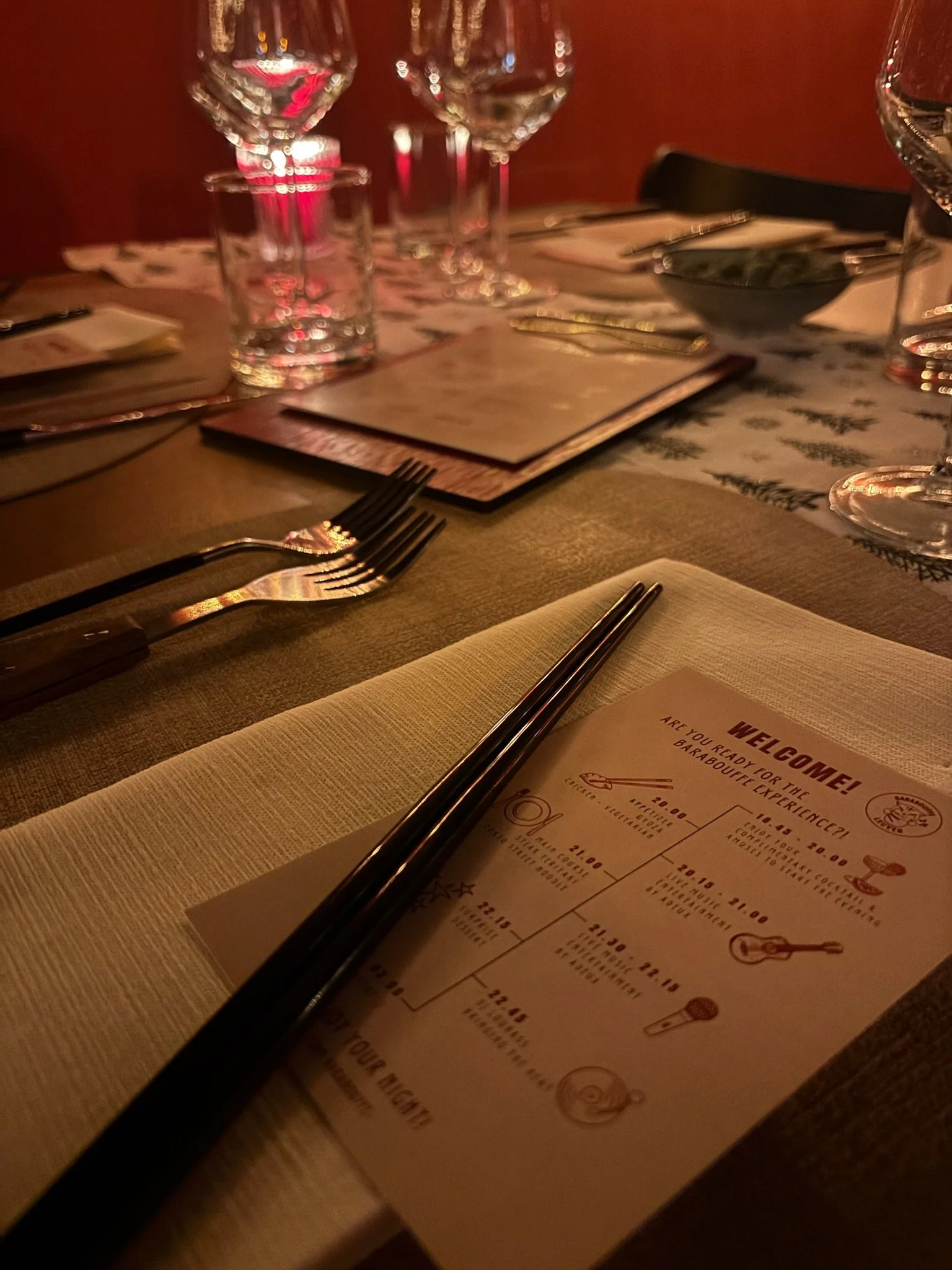 Elegant restaurant table set with wine glasses, water glasses, silverware, menu, and chopsticks on a beige napkin under warm lighting.