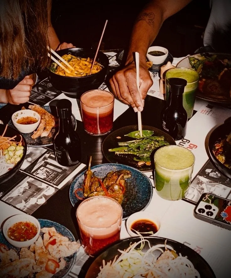 A table with various dishes of Asian cuisine, drinks, and people sharing a meal at a restaurant.