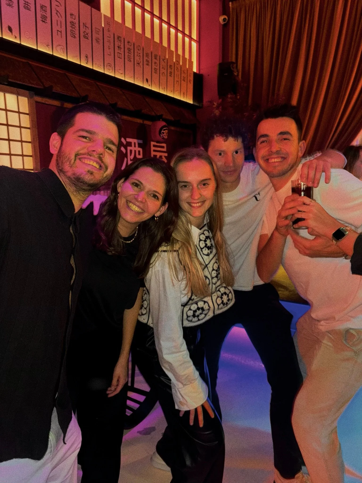A group of five friends smiling and posing together at a bar or nightclub, holding drinks, with colorful lighting and Asian-themed decor in the background.