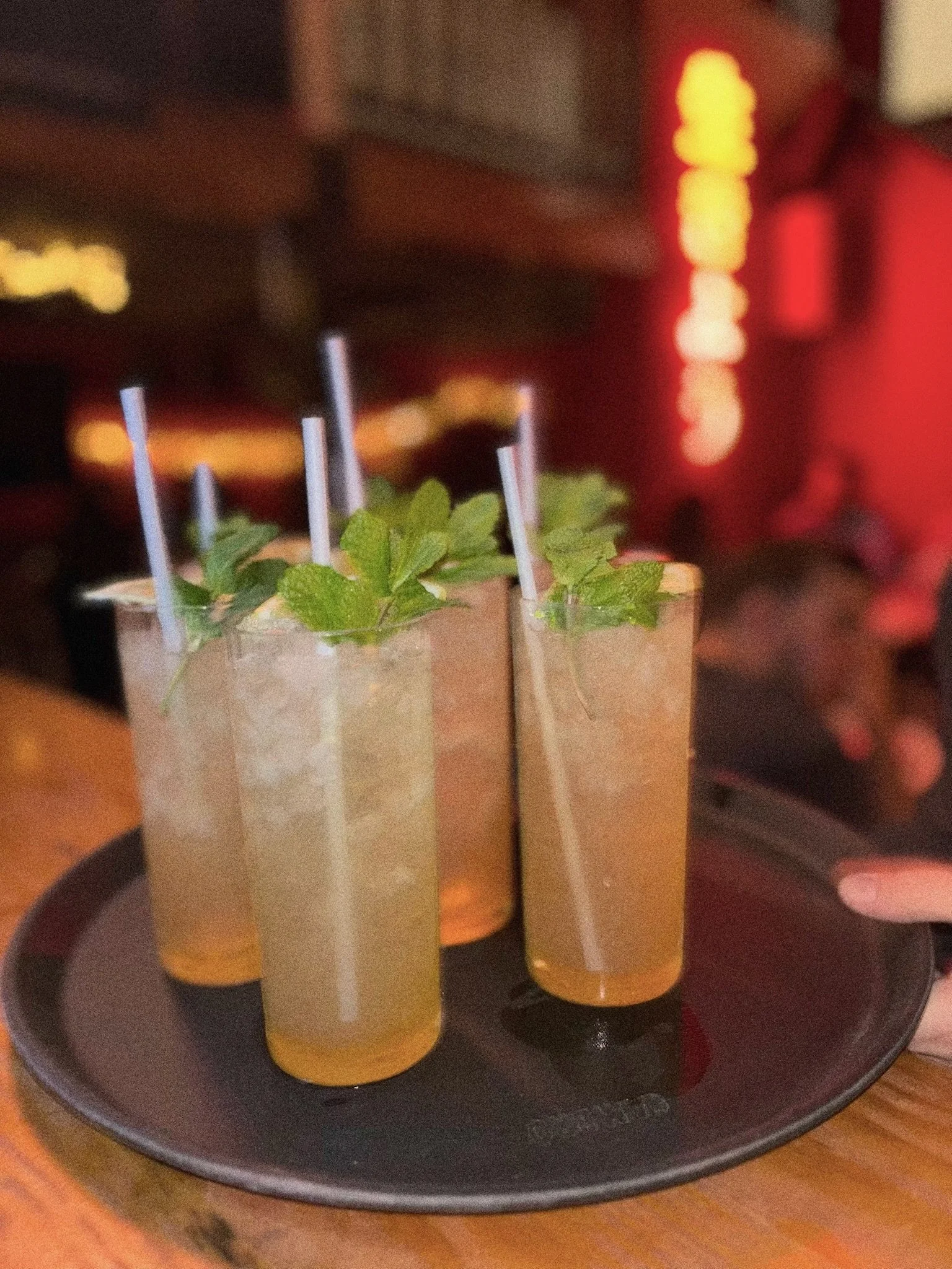 Four tall glasses of a light-colored drink garnished with mint leaves and straws, served on a black tray in a dimly lit setting.