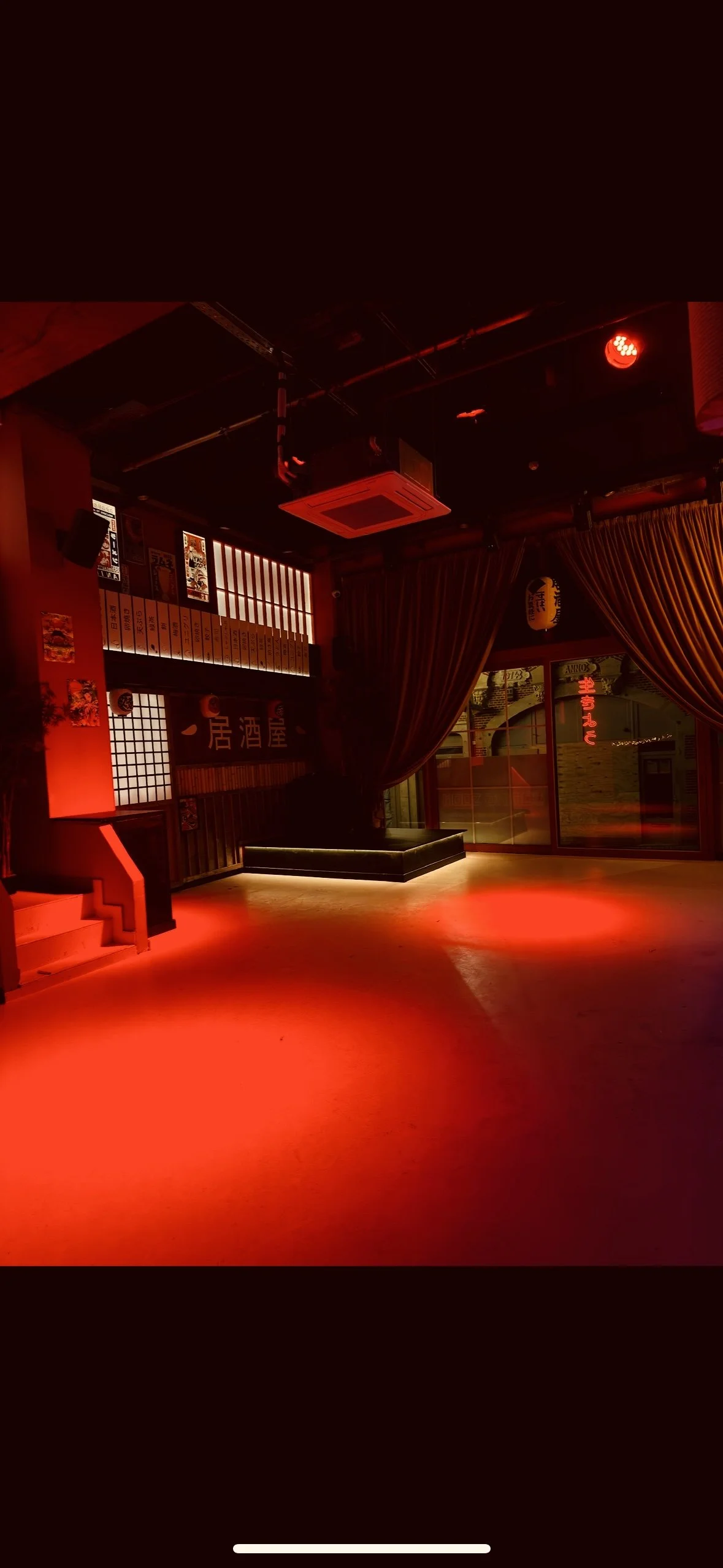 Empty Japanese-style bar with red lighting, wooden decor, a small stage area, and closed curtains.