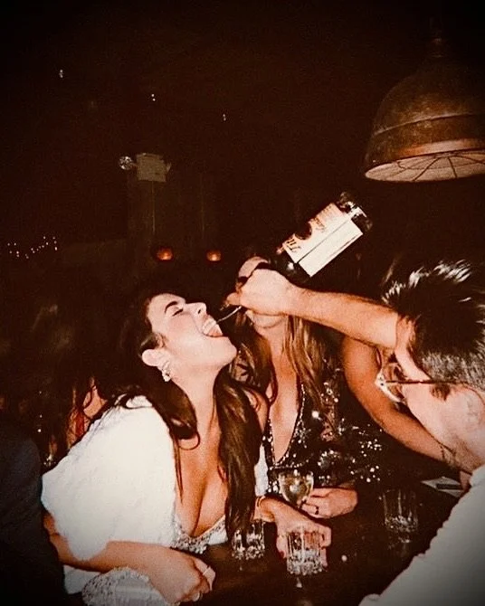 A woman at a bar is being shot with a liquor bottle by another woman, both smiling and having fun.