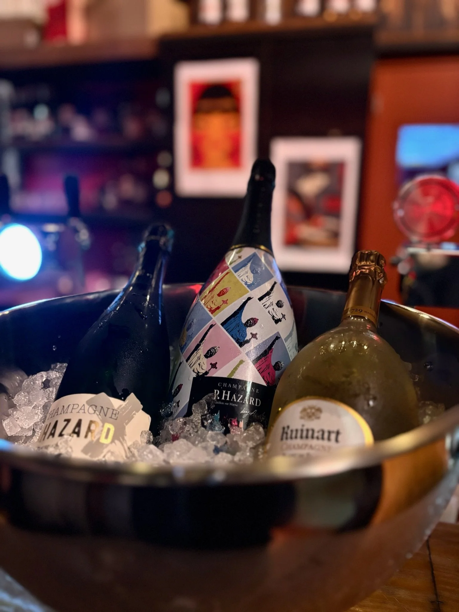 Three bottles of champagne in a metal ice bucket filled with ice, with blurred artwork on the wall in the background.