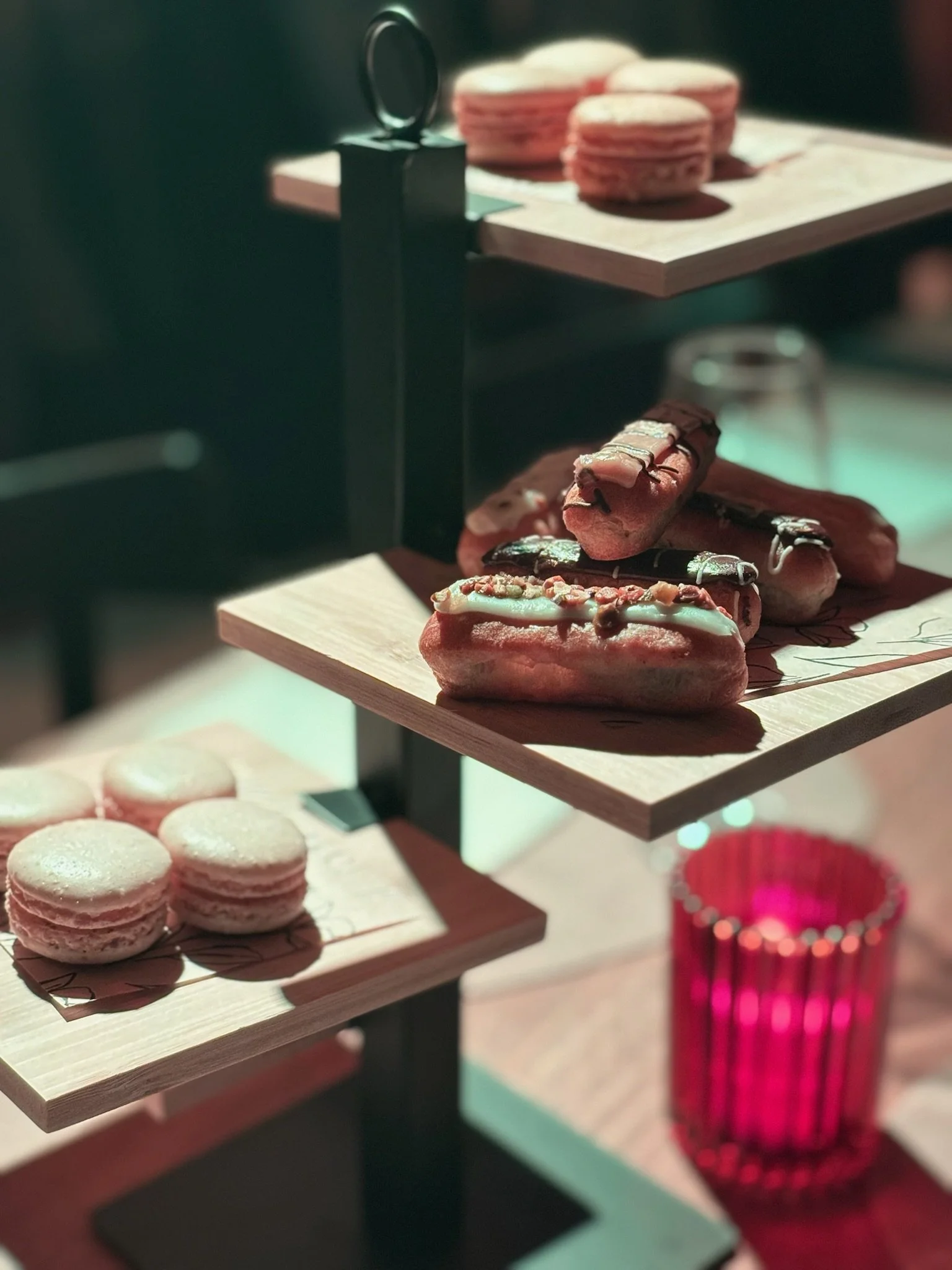 Assorted macarons and decorated donuts displayed on a multi-tiered wooden stand in a cafe or bakery setting.