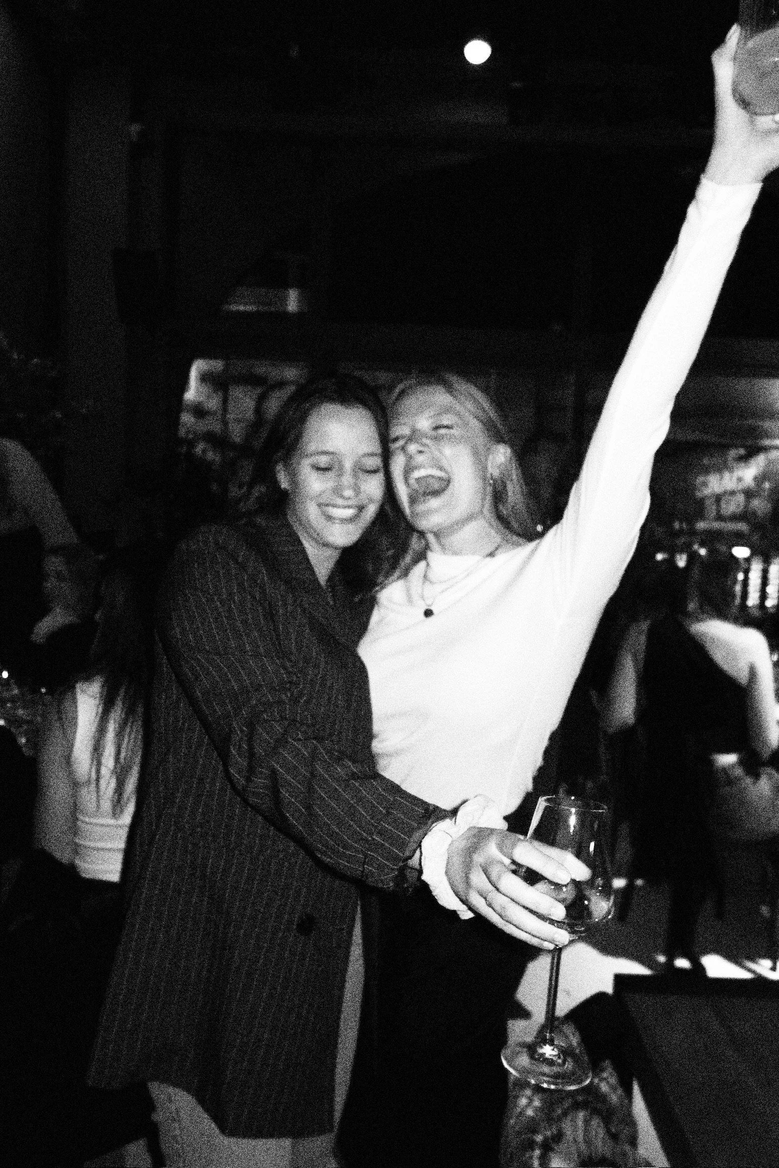Two women smiling and celebrating, one holding a glass of wine, at a social gathering or party.