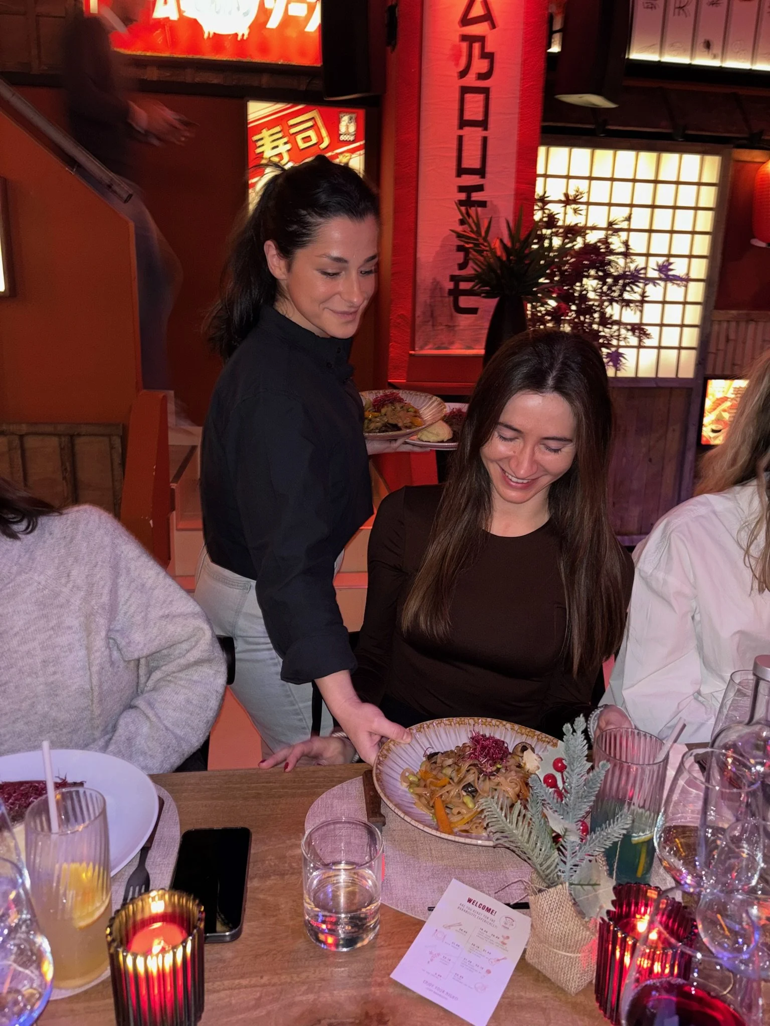 Two women at a restaurant table with food and drinks, one serving pasta to the other, with Asian-themed decor and red lanterns in the background.