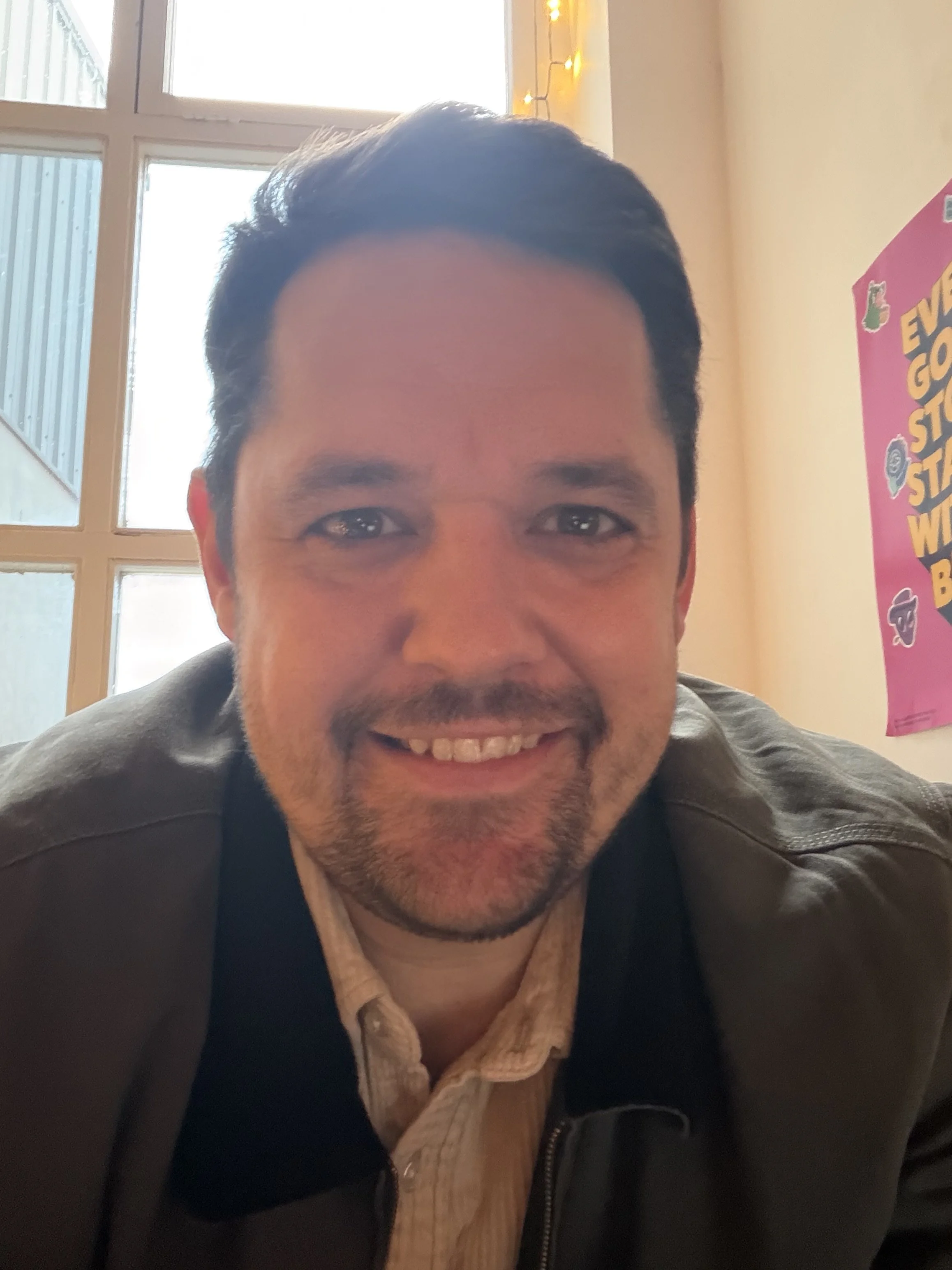 A smiling man with dark hair and facial hair, wearing a jacket and a collared shirt, in a room with windows and string lights.