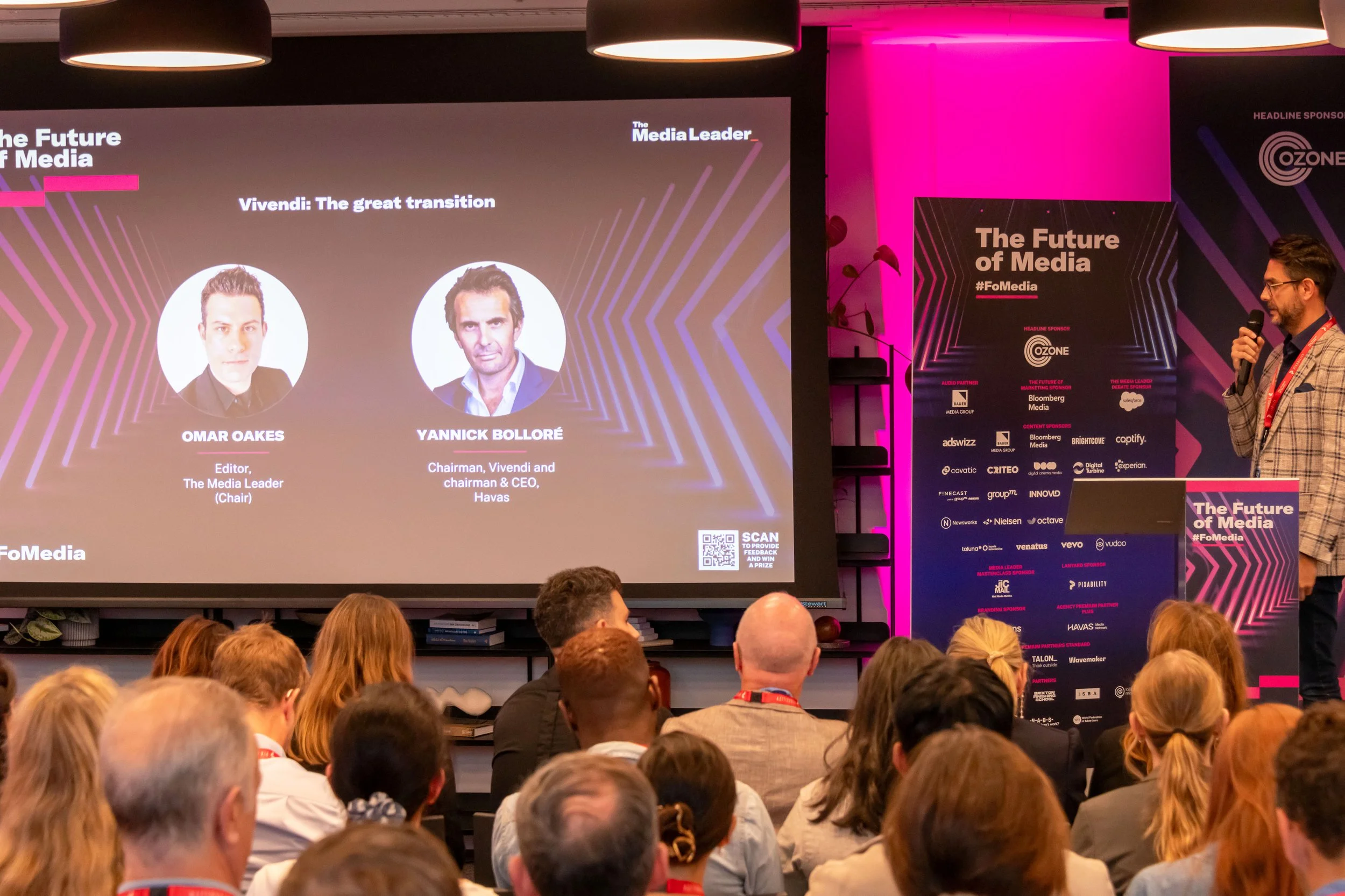 Presentation at a conference titled 'The Future of Media', showing two speakers on a large screen with their names and titles: Omar Oakes, Editor of The Media Leader, and Yannick Bolloré, Chairman of Vivendi and CEO of Havas. An audience watches, and a man in a checked blazer speaks into a microphone at the podium with the conference branding.