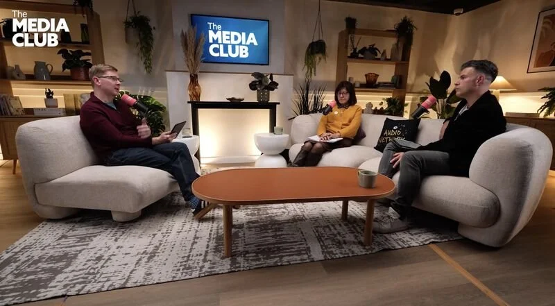 Three people sitting on couches in a studio set of 'The Media Club,' engaged in a discussion. Two men and one woman, with the man on the left holding a microphone and some papers, the woman in the middle taking notes, and the man on the right with a microphone. A TV screen displaying the show's logo is in the background, with plants and shelves decor on the set.