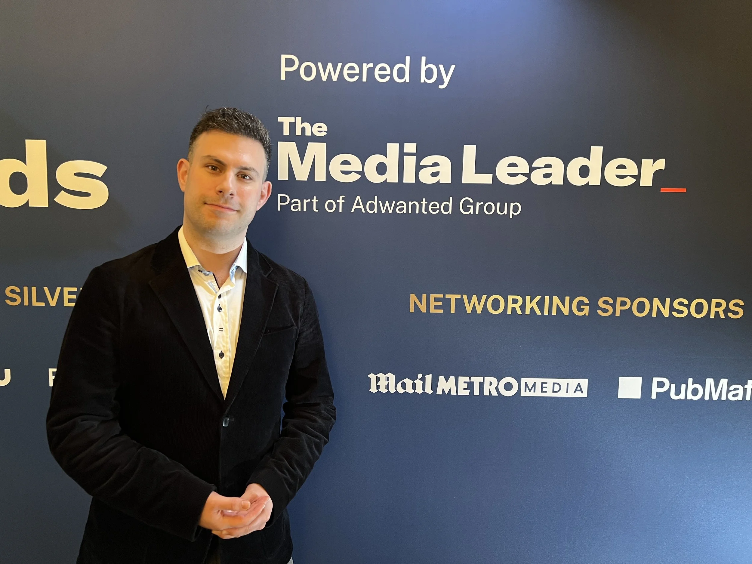 A man in a black blazer and a patterned white shirt standing in front of a blue backdrop with text, including "Powered by The Media Leader," and logos for Mail Metro Media and PubMatic.