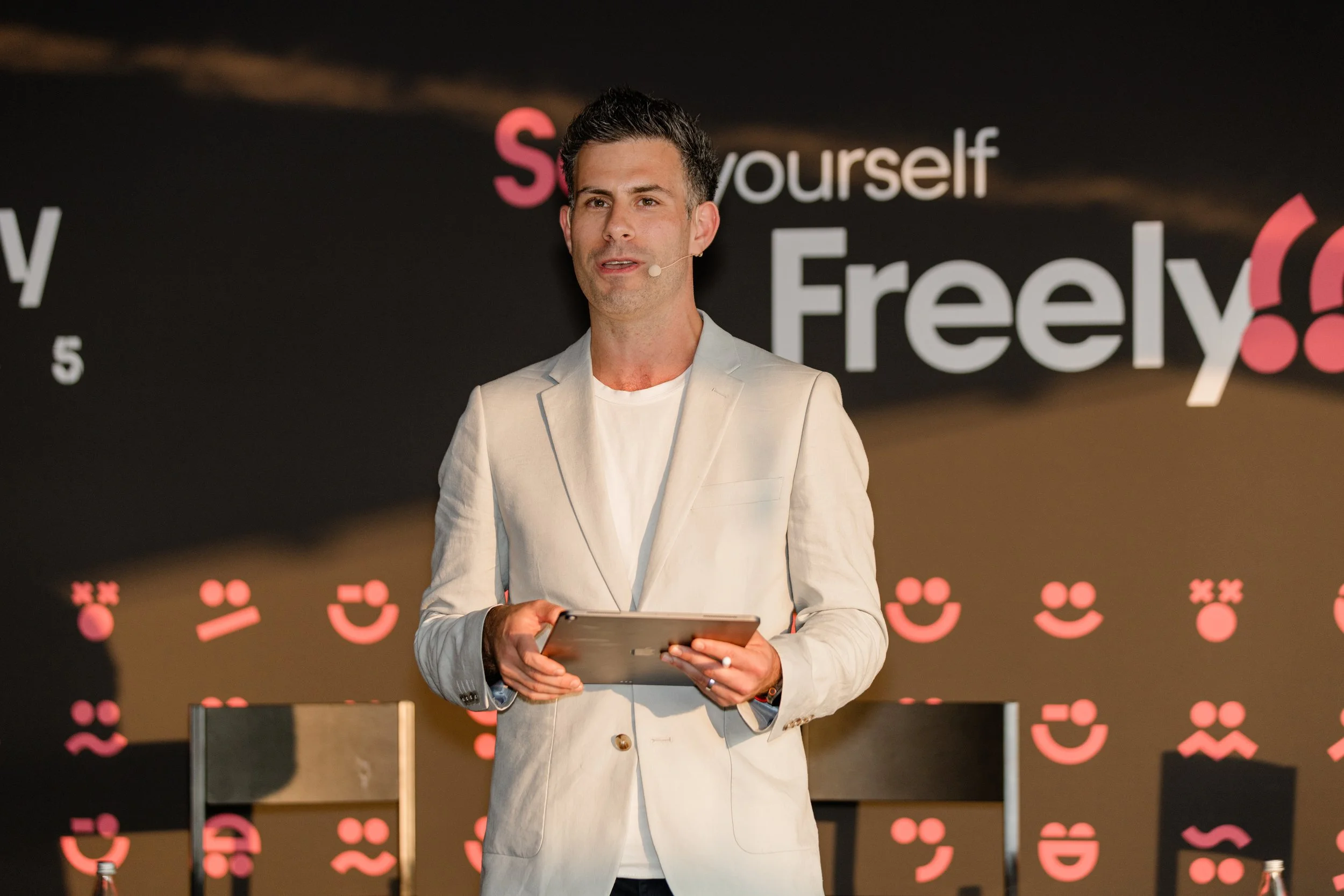 Man in light-colored blazer holding a tablet and speaking at a presentation with a black background, white and pink text, and pink emoticons.