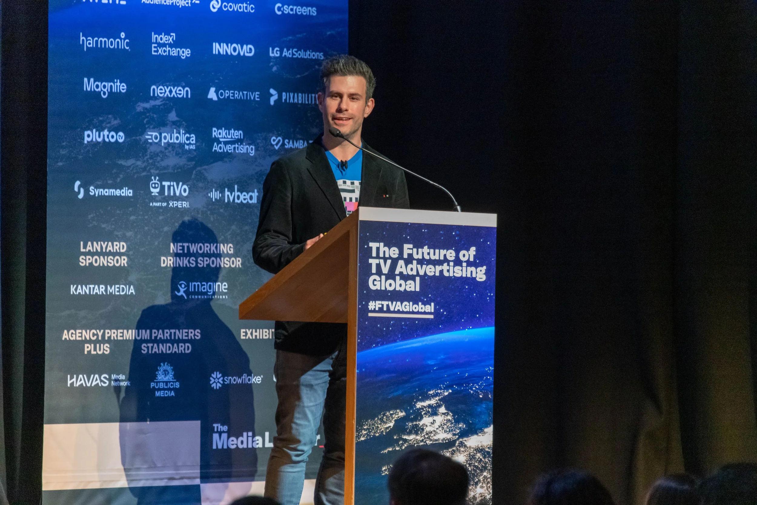 A man standing at a podium giving a presentation at a conference titled 'The Future of TV Advertising Global'. The background features a screen displaying logos of various sponsors and partners.