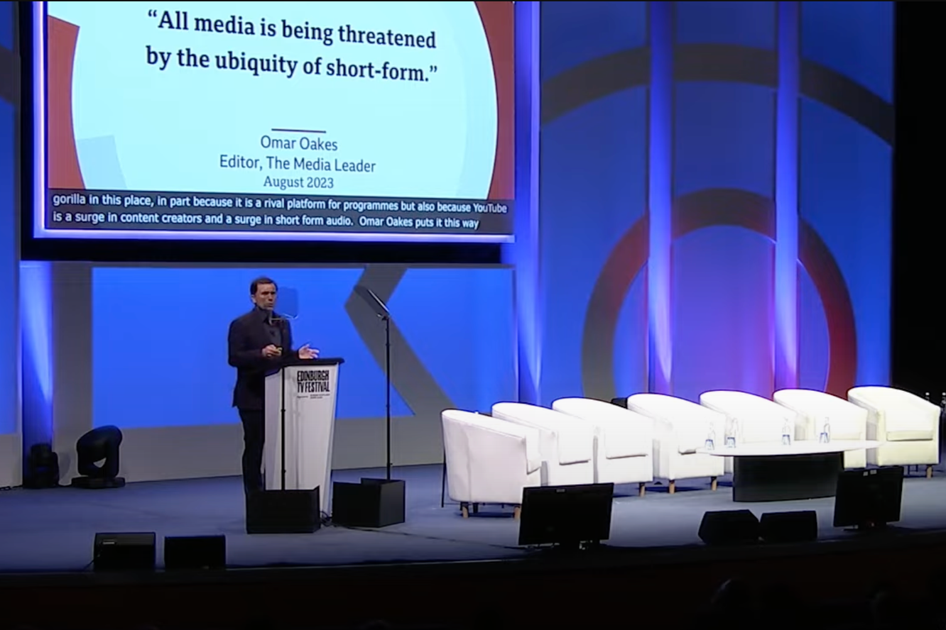 A man in a dark suit speaking at a podium on a stage with blue lighting. Behind him, there are six white chairs and a large screen displaying a quote: 'All media is being threatened by the ubiquity of short-form,' attributed to Omar Oakes, August 2023. The stage is part of an event called the Edinburgh TV Festival.