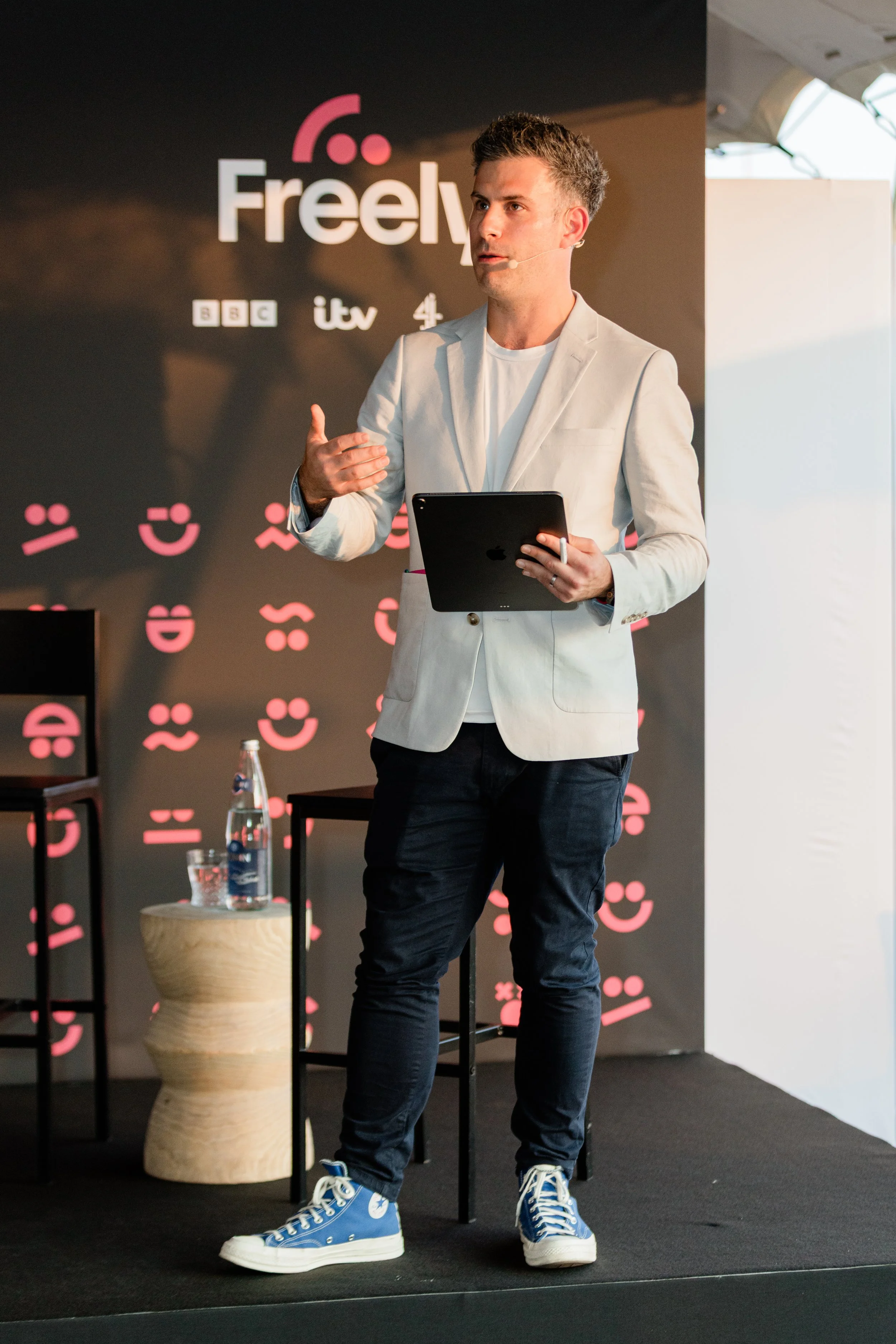 A man is speaking on stage at an event, holding a tablet and gesturing with his hand. He is wearing a white blazer, black pants, and blue high-top sneakers. The background has the word 'Freely' and logos of BBC, ITV, and 4.