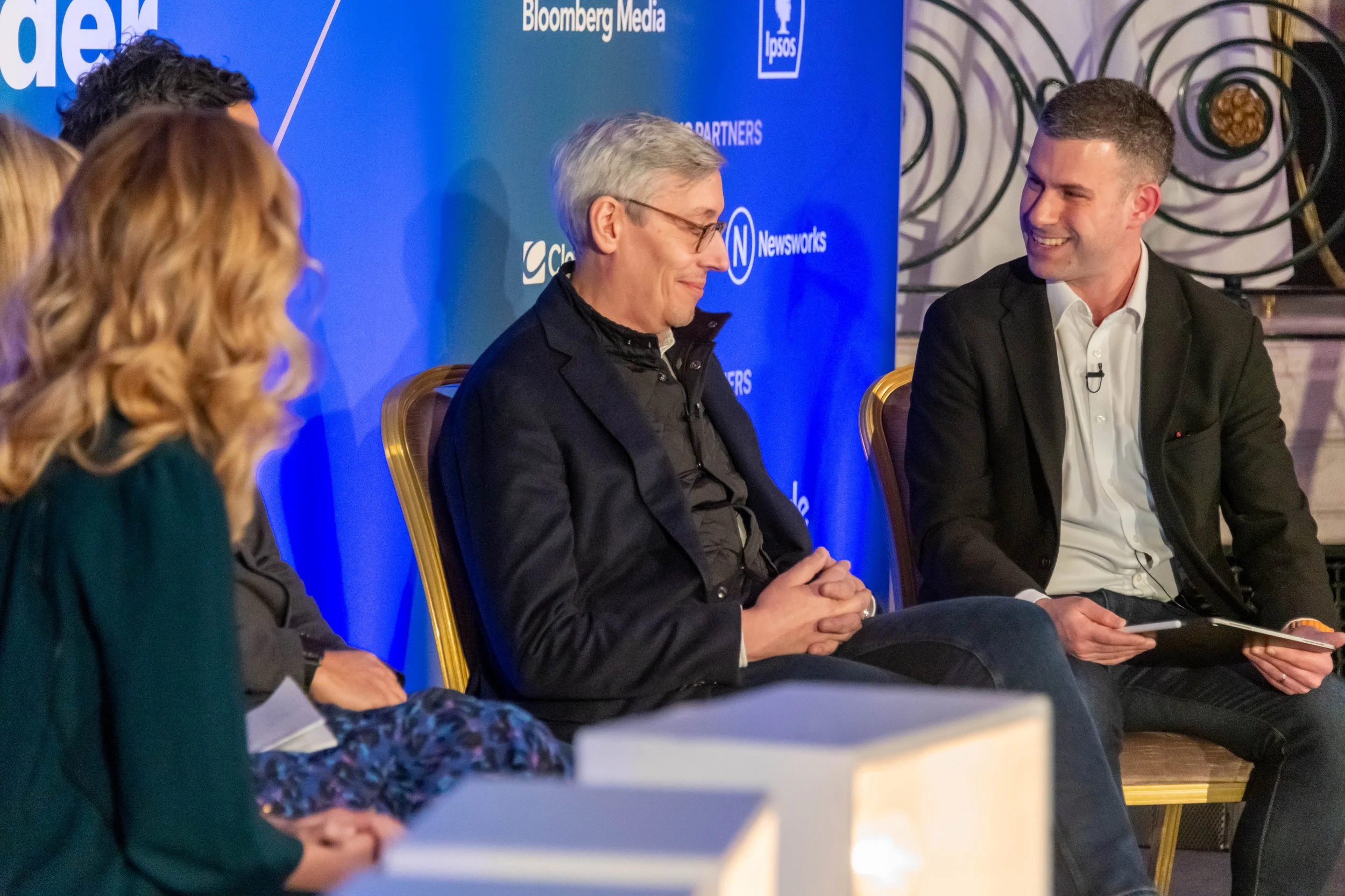 A panel of four people seated on stage during a discussion, with a blue backdrop displaying logos including Bloomberg Media, Newsworks, and Lipos.