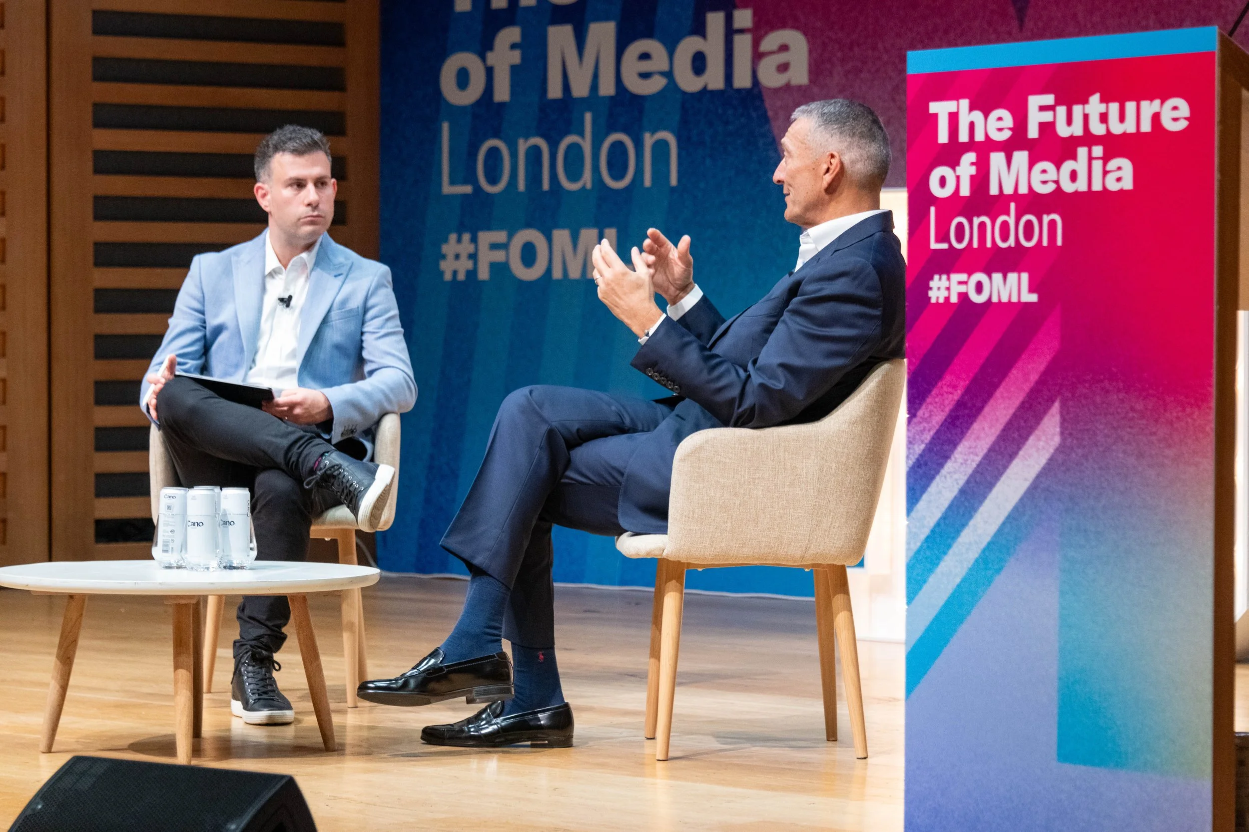 Two men seated on a stage engaged in a conversation at a media event. One man is in a light blue blazer and the other in a dark suit. A backdrop displays the text 'The Future of Media London #FOML', and there is a side banner with similar text.