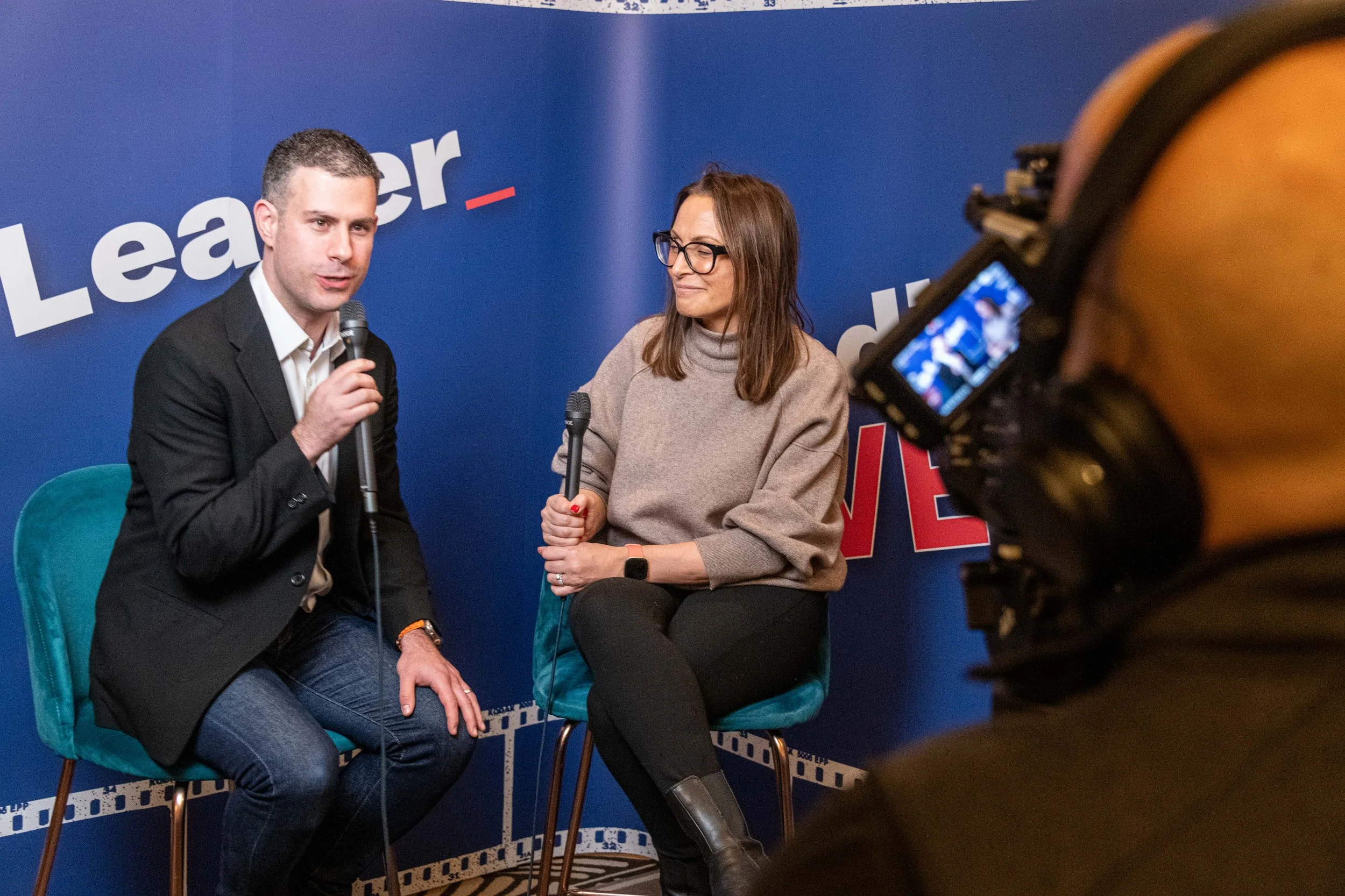 A man and a woman sitting on chairs during an interview or discussion, with a camera operator recording them, and a blue backdrop with partially visible text.