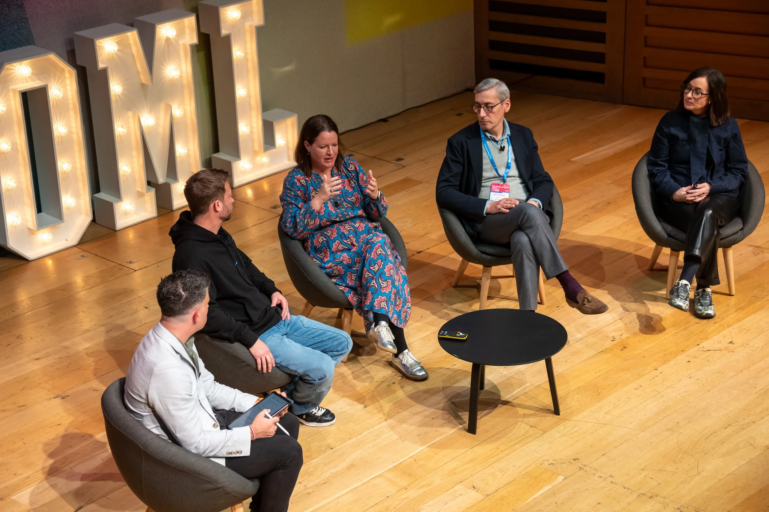 A panel discussion with five people on a stage. Four are seated in chairs, one in a white jacket with a tablet. Large illuminated letters spell 'OMI' in the background.