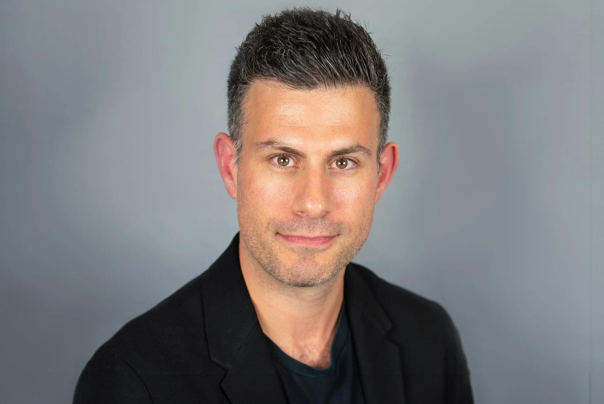 Headshot of a man with short dark hair, light skin, wearing a black blazer and black shirt, smiling slightly against a plain grey background.