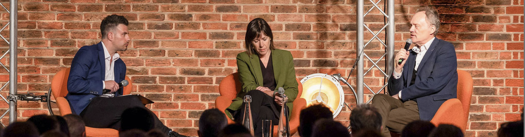 Three people engaging in a panel discussion or interview on stage, seated in orange chairs in front of a brick wall, with a small audience watching.