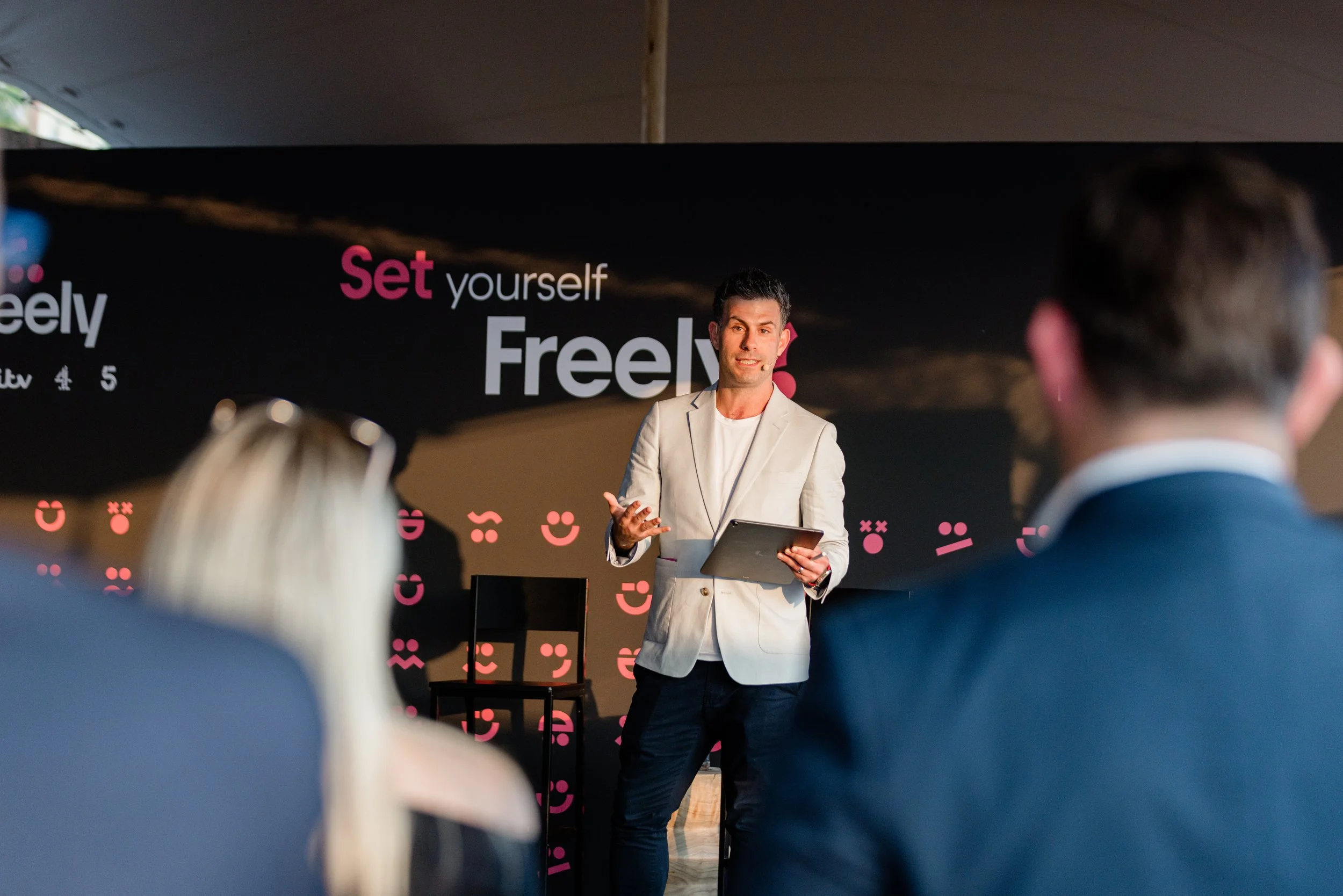 Man in a white blazer giving a presentation to an audience with a black backdrop and pink text that reads 'Set yourself Free'.