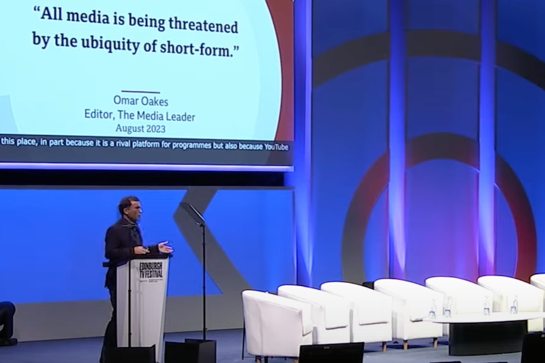 A man standing at a podium on a stage giving a presentation. A large screen behind him displays a quote: 'All media is being threatened by the ubiquity of short-form.' The quote is attributed to Omar Oakes, Editor of The Media Leader, August 2023. The stage has white chairs and a colorful background.