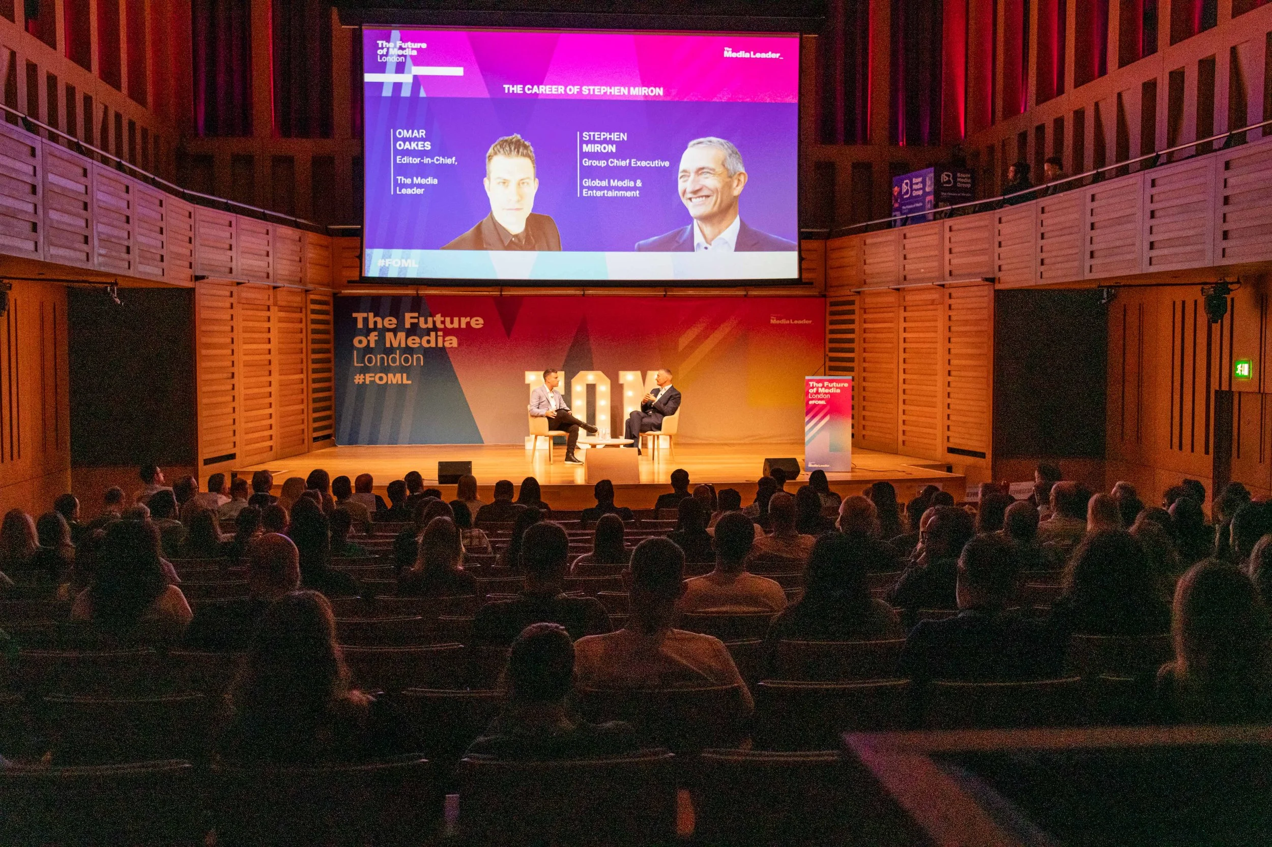 A stage at an event called The Future of Media London, #FOML, with two men seated on stage in conversation. One man is holding a microphone, and the background has a large screen with images and names of Omar Oakes and Stephen Miron.