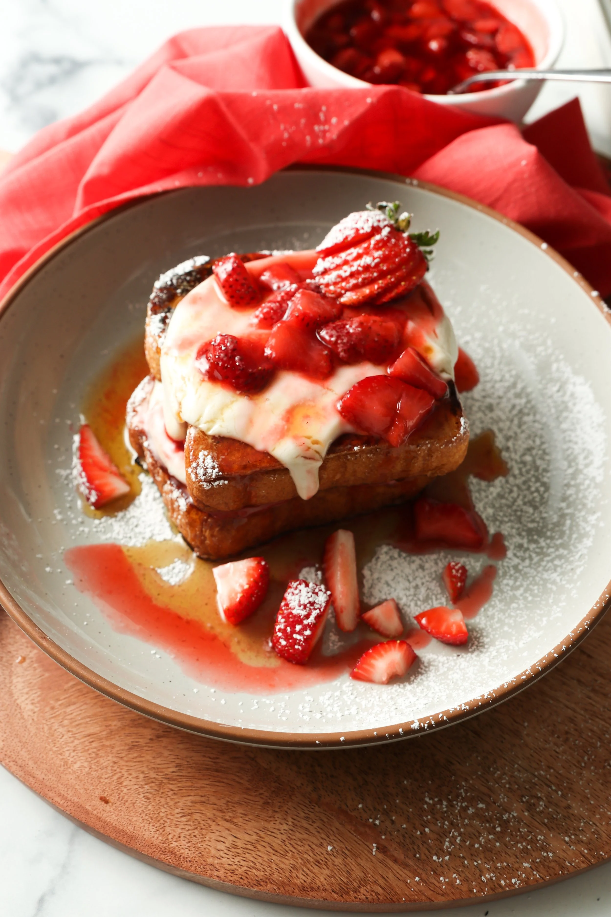 French toast topped with cream cheese frosting, strawberries, and syrup on a white plate, with additional strawberries and syrup in a bowl in the background.