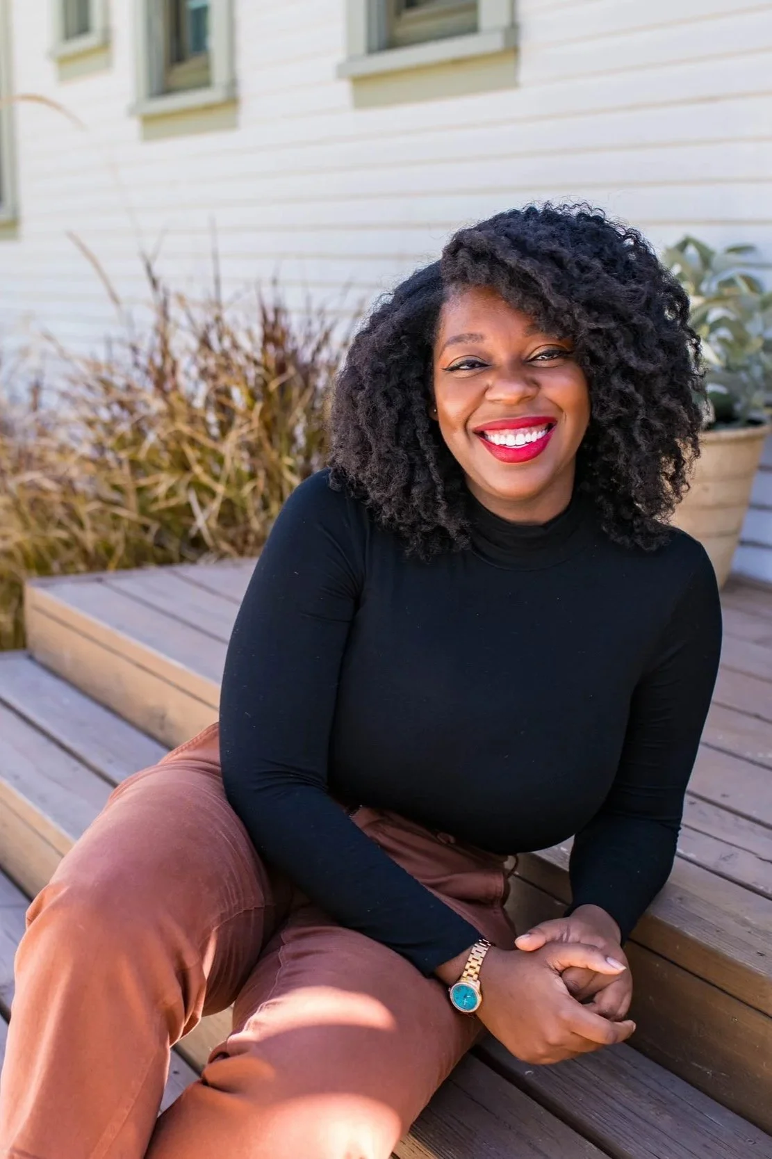 A woman with curly hair wearing a black turtleneck and brown pants, sitting on wooden steps outside, smiling with red lipstick, near a potted plant and leafless bushes.