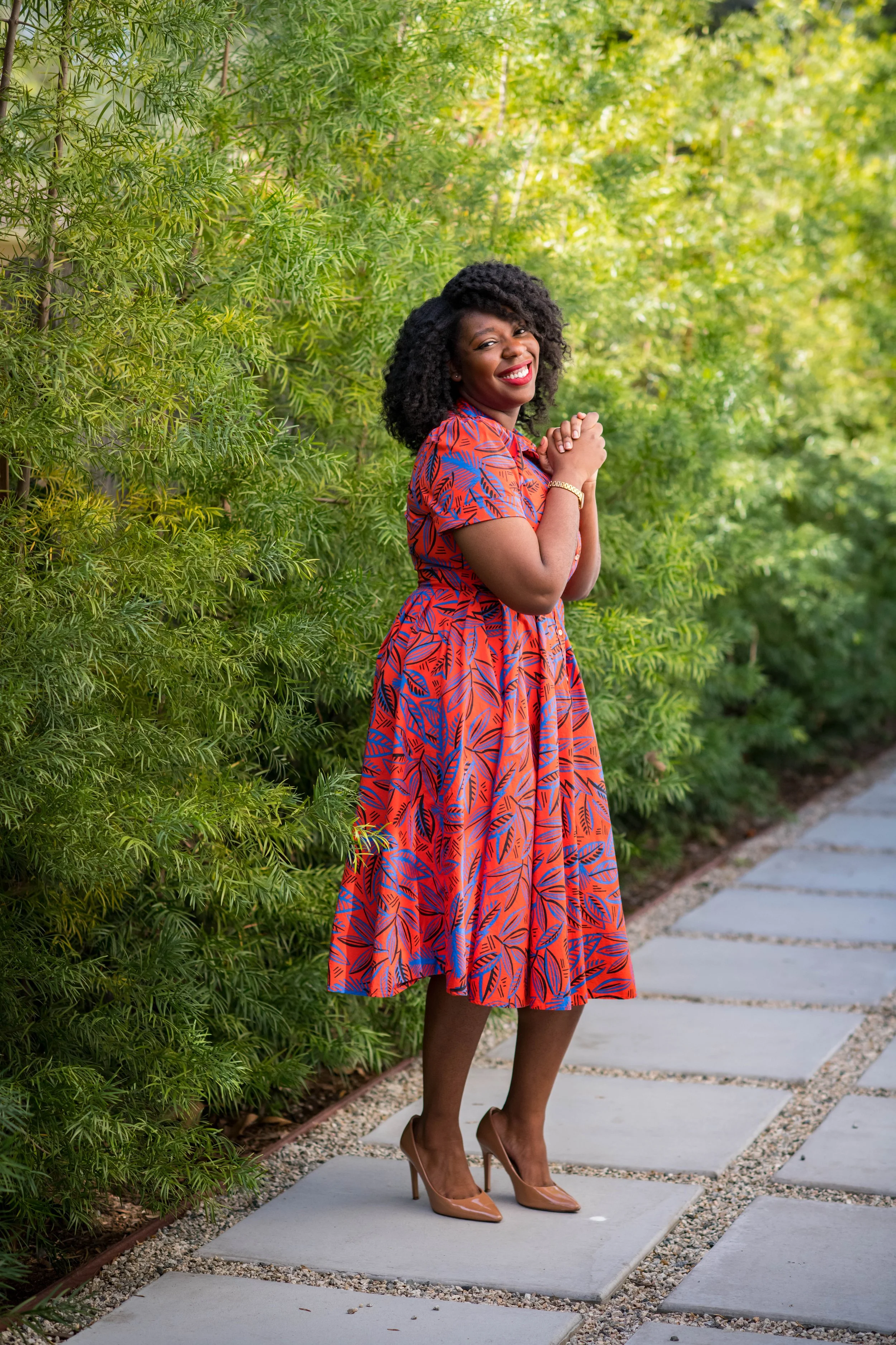 A smiling woman with curly hair in a colorful dress stands on a stone pathway, surrounded by green bushes.