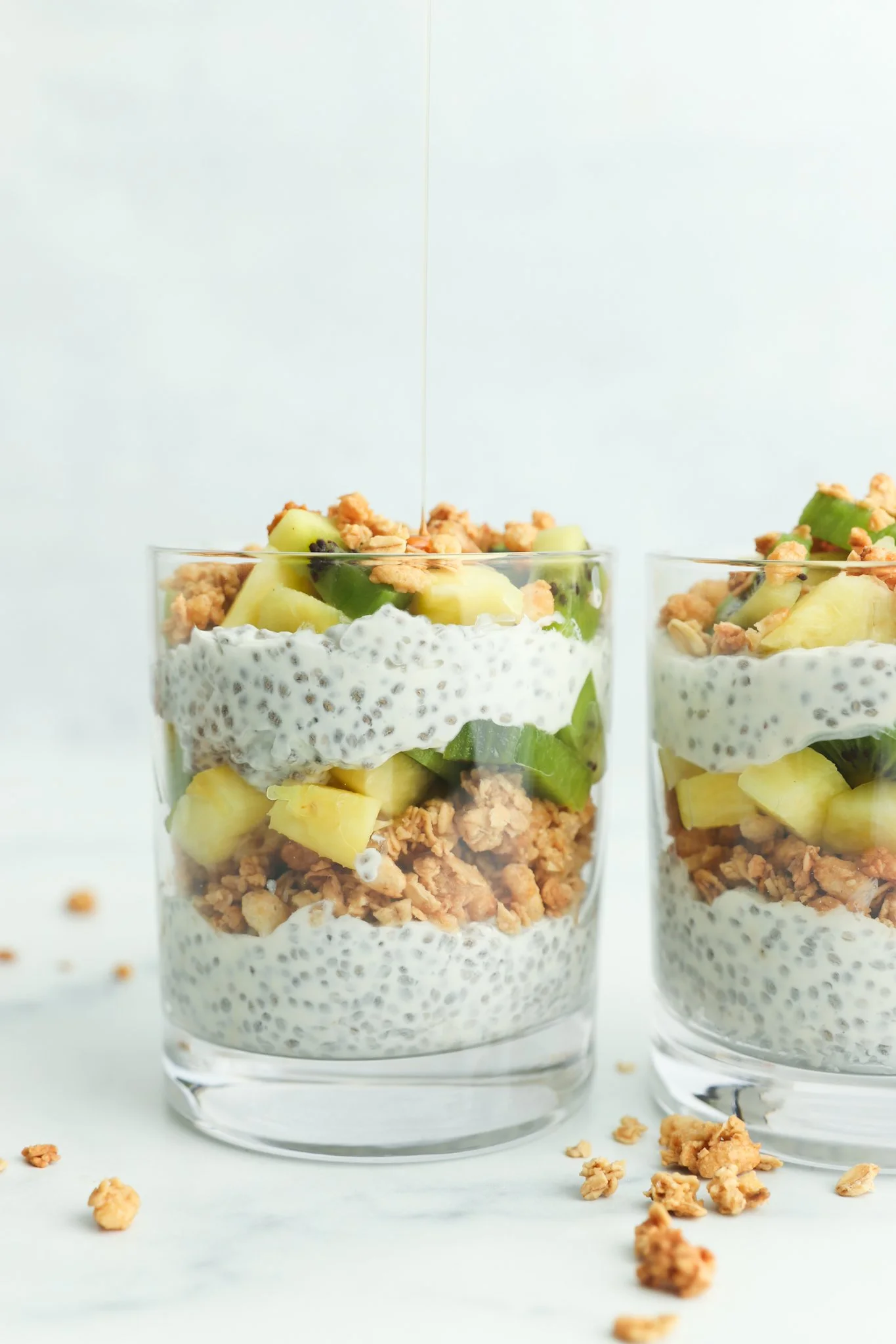 Two glasses of chia seed pudding layered with granola, pineapple, and kiwi, with honey being drizzled on top.