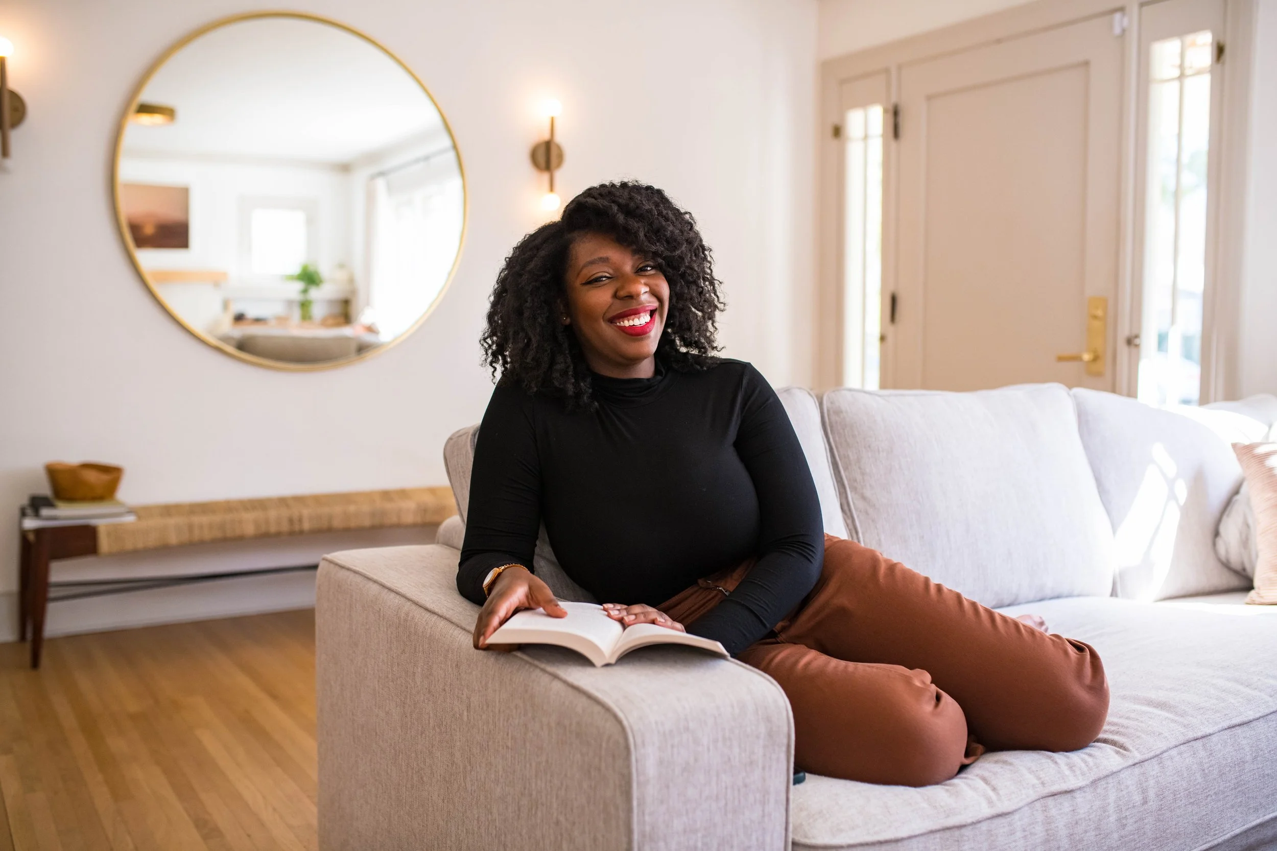 A woman with dark curly hair, wearing a black turtleneck and brown pants, sitting on a light-colored sofa in a bright living room, smiling and holding an open book in her lap.