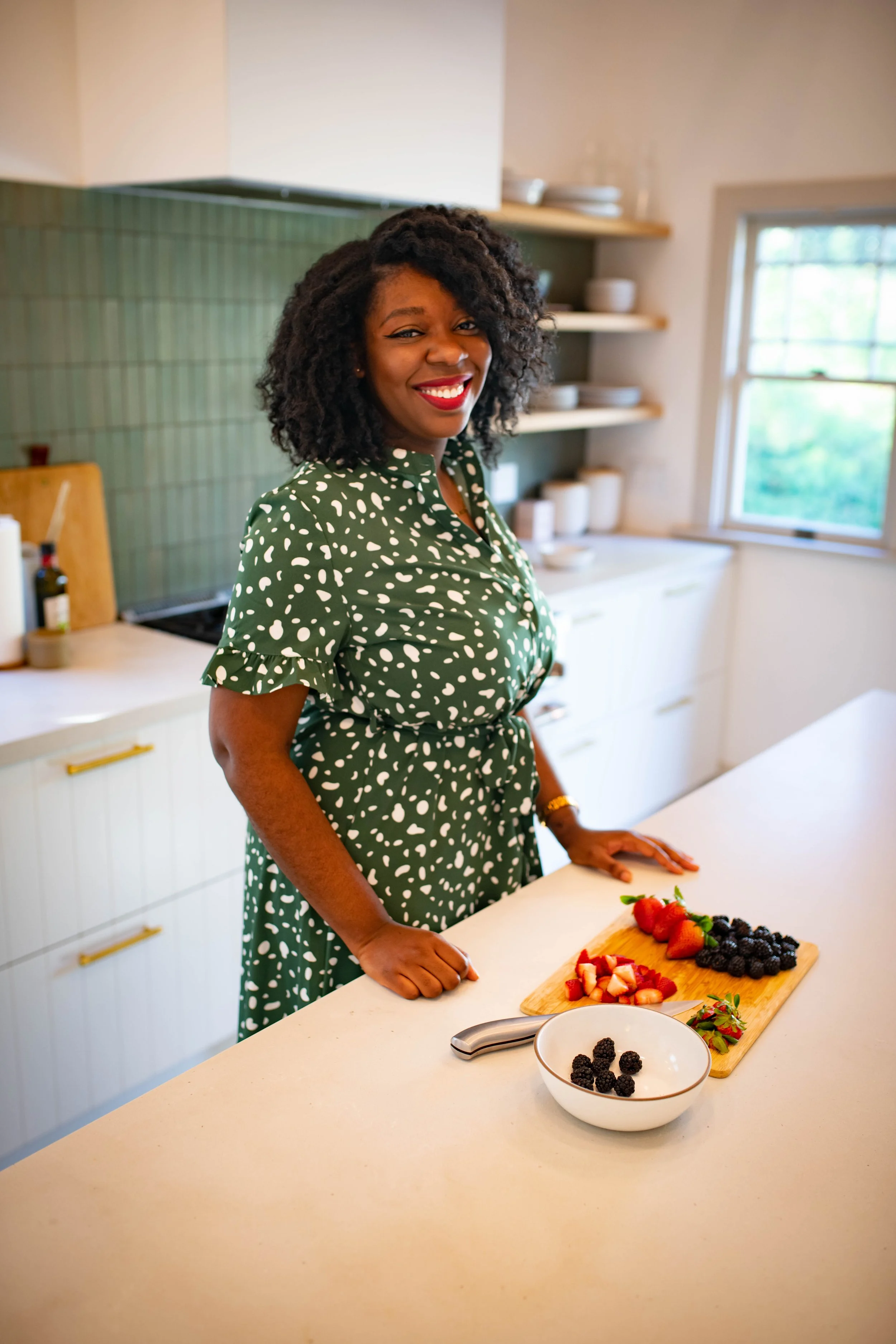 A woman with curly hair and red lipstick smiling in a kitchen, standing behind a white counter with a chopping board of strawberries, blackberries, and cut strawberries, and a bowl of blackberries.