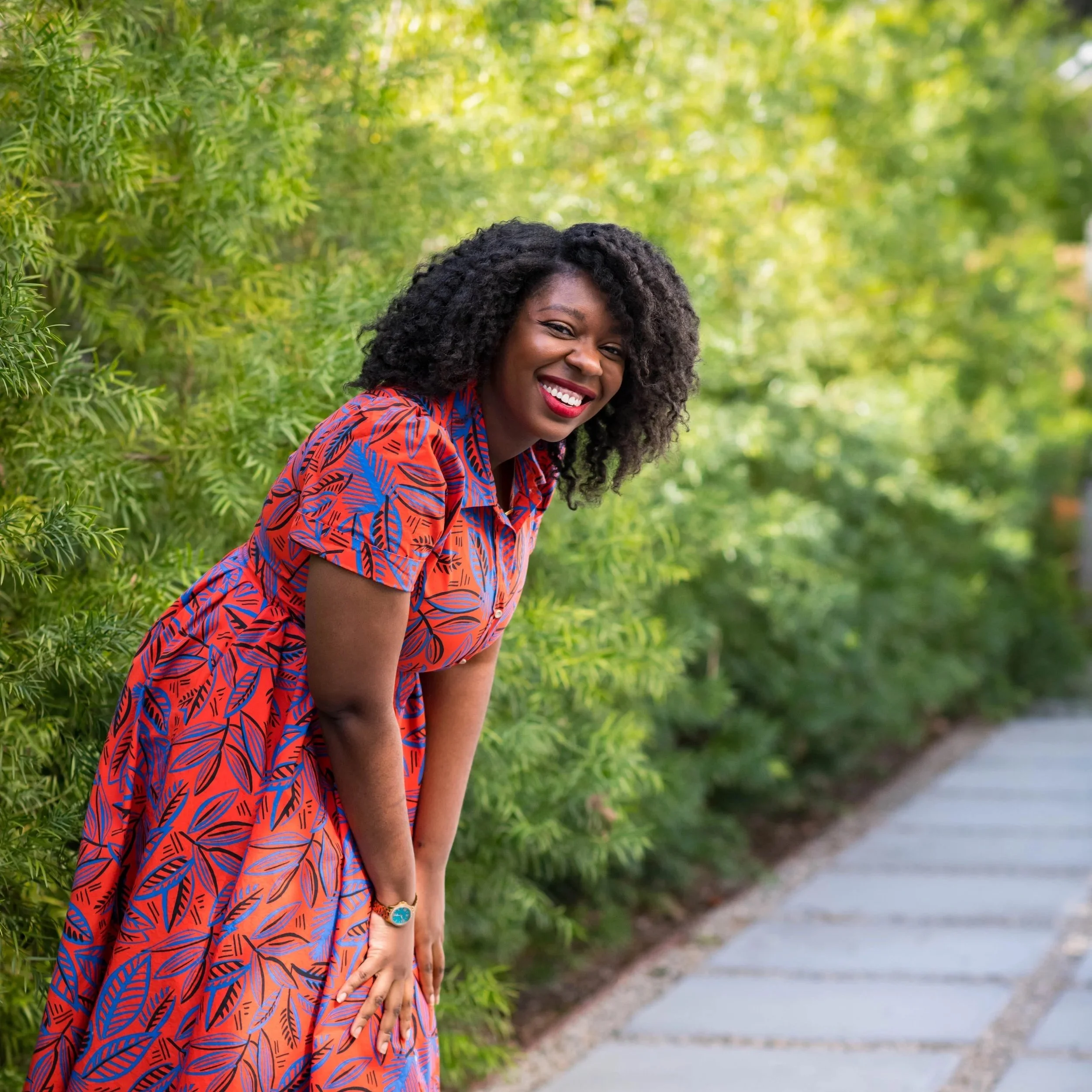 A woman with curly black hair, wearing a colorful patterned dress, smiling and bending forward outdoors on a sidewalk with green bushes in the background.