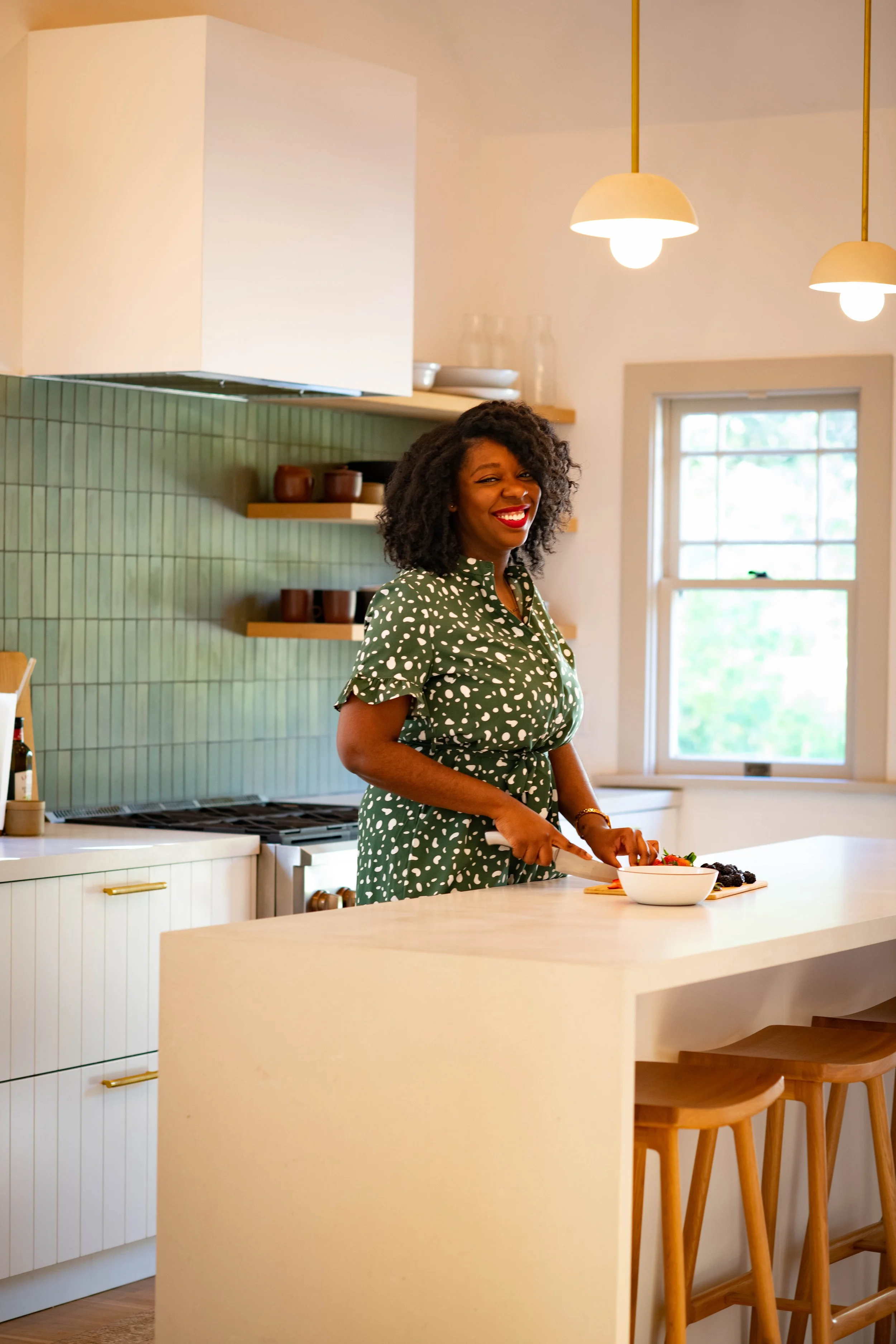 A woman with curly hair, wearing a green dress with white spots, is smiling while slicing fruit in a bright, modern kitchen with a white island and wooden bar stools.
