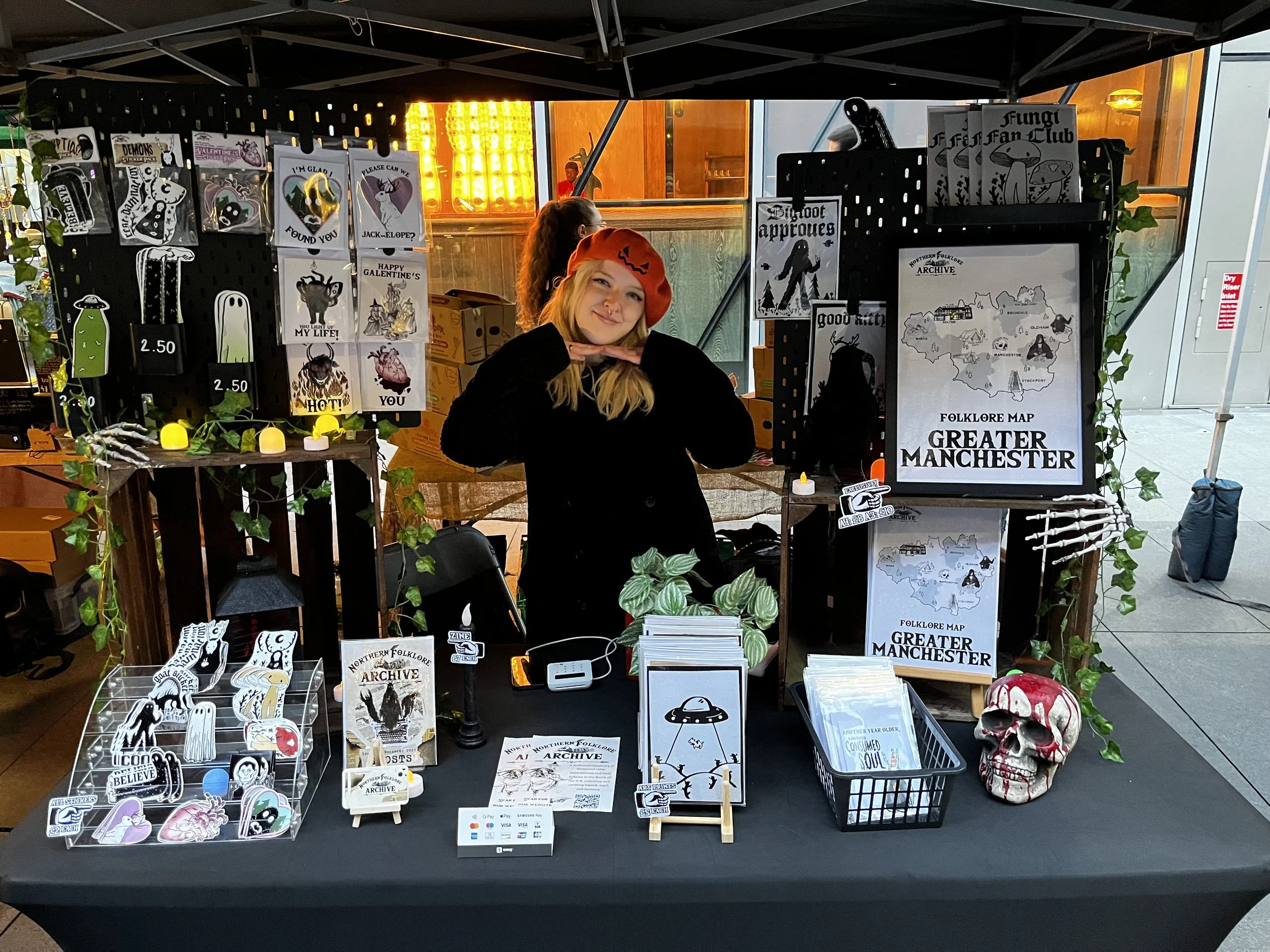 A woman standing behind a booth at a market, displaying various merchandise such as postcards, stickers, a folklore map of Greater Manchester, and a decorative skull with red accents. The booth is decorated with green vines and small yellow lights.